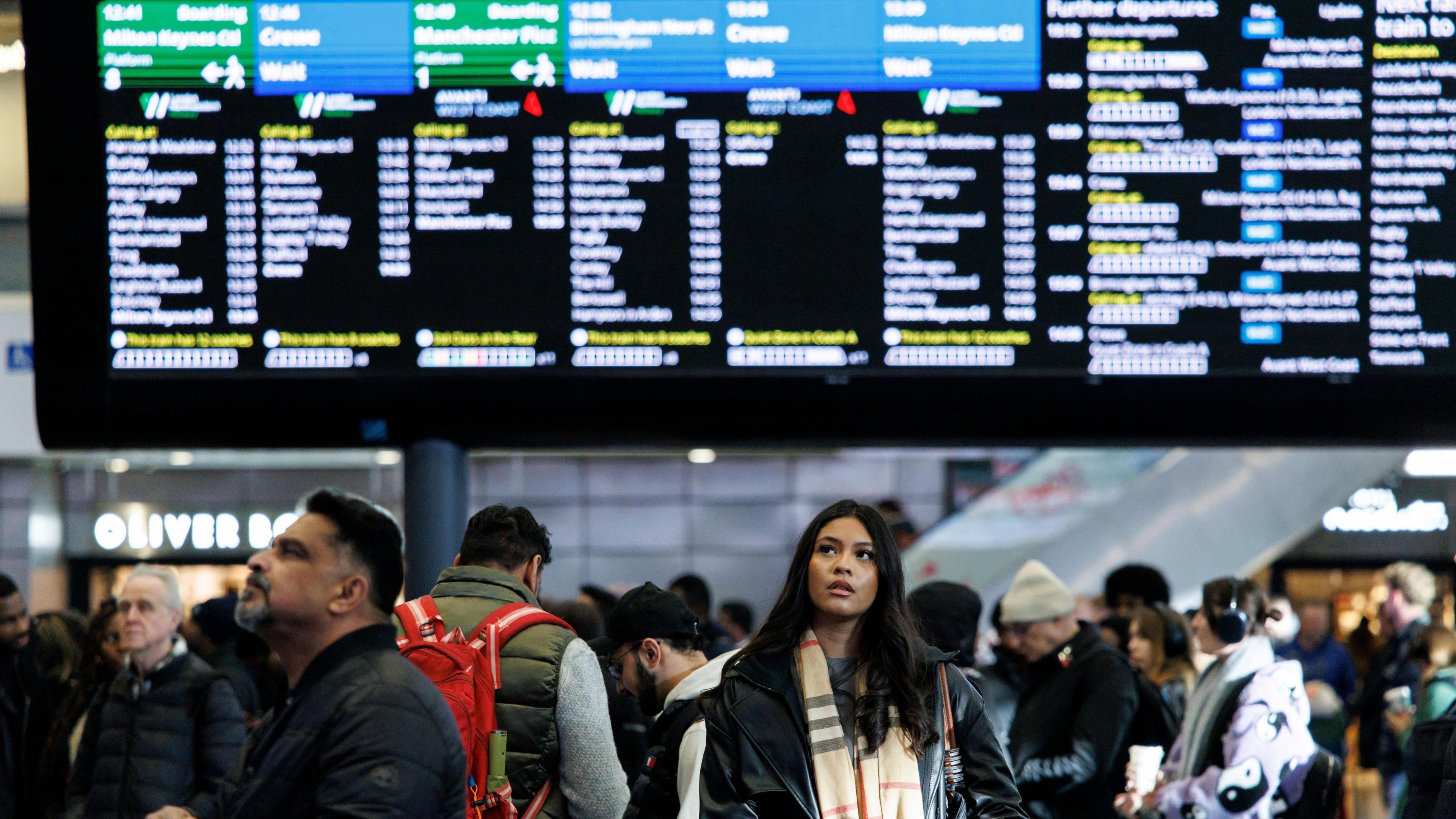 A woman with long-dark hair and wearing a beige-patterned scarf stands looking up under electronic departure boards at a busy Euston Station