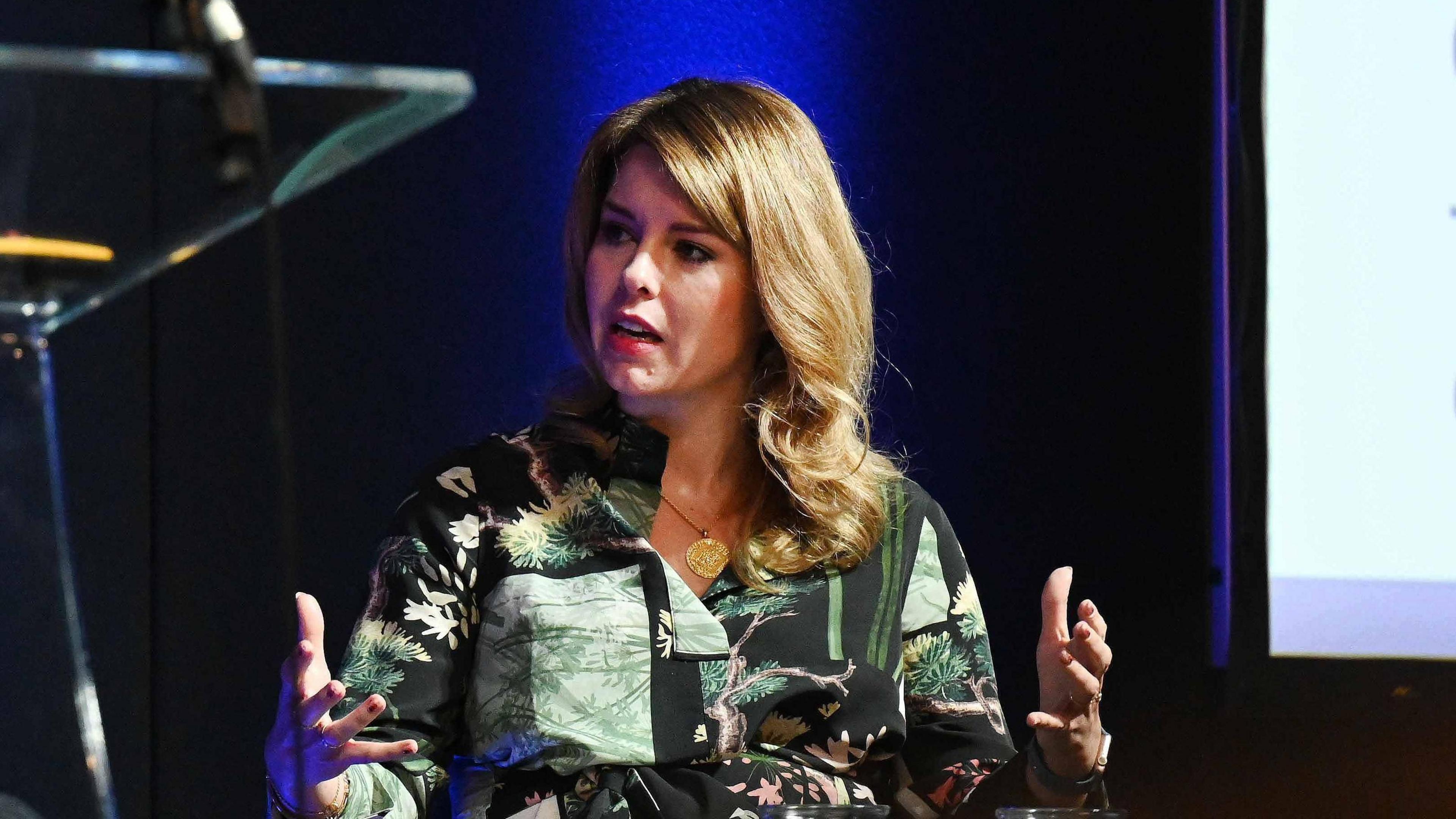 Kim McGuinness sitting on a chair with her hands in the air. She is wearing a patterned green top of tree images and has blonde hair.