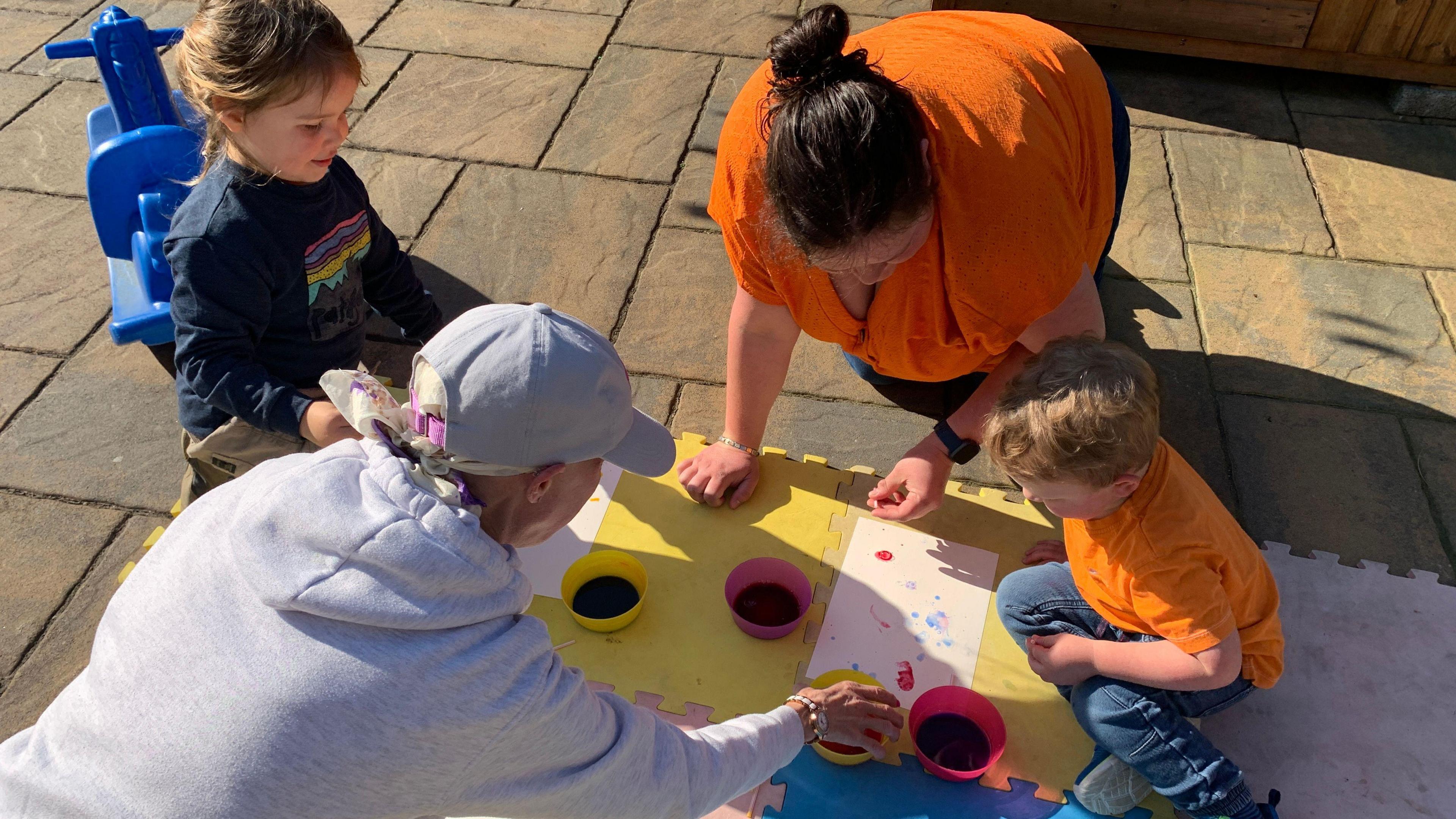 Two children lie on the ground playing with finger paints. The adults - mum Rebecca and childminder Suzi help them