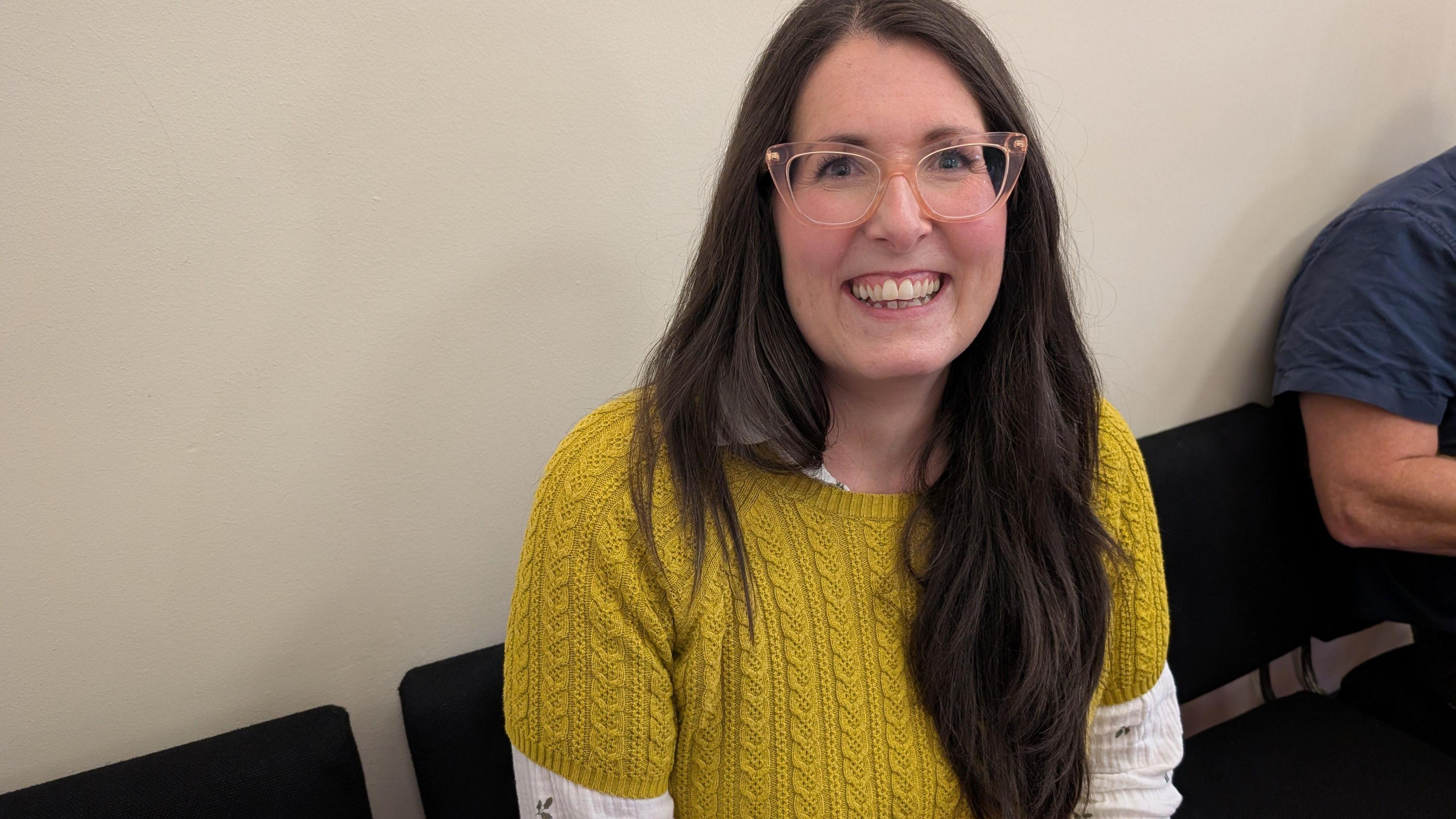 A woman wearing a yellow jumper with white sleeves sits on a chair, whilst smiling into the camera.