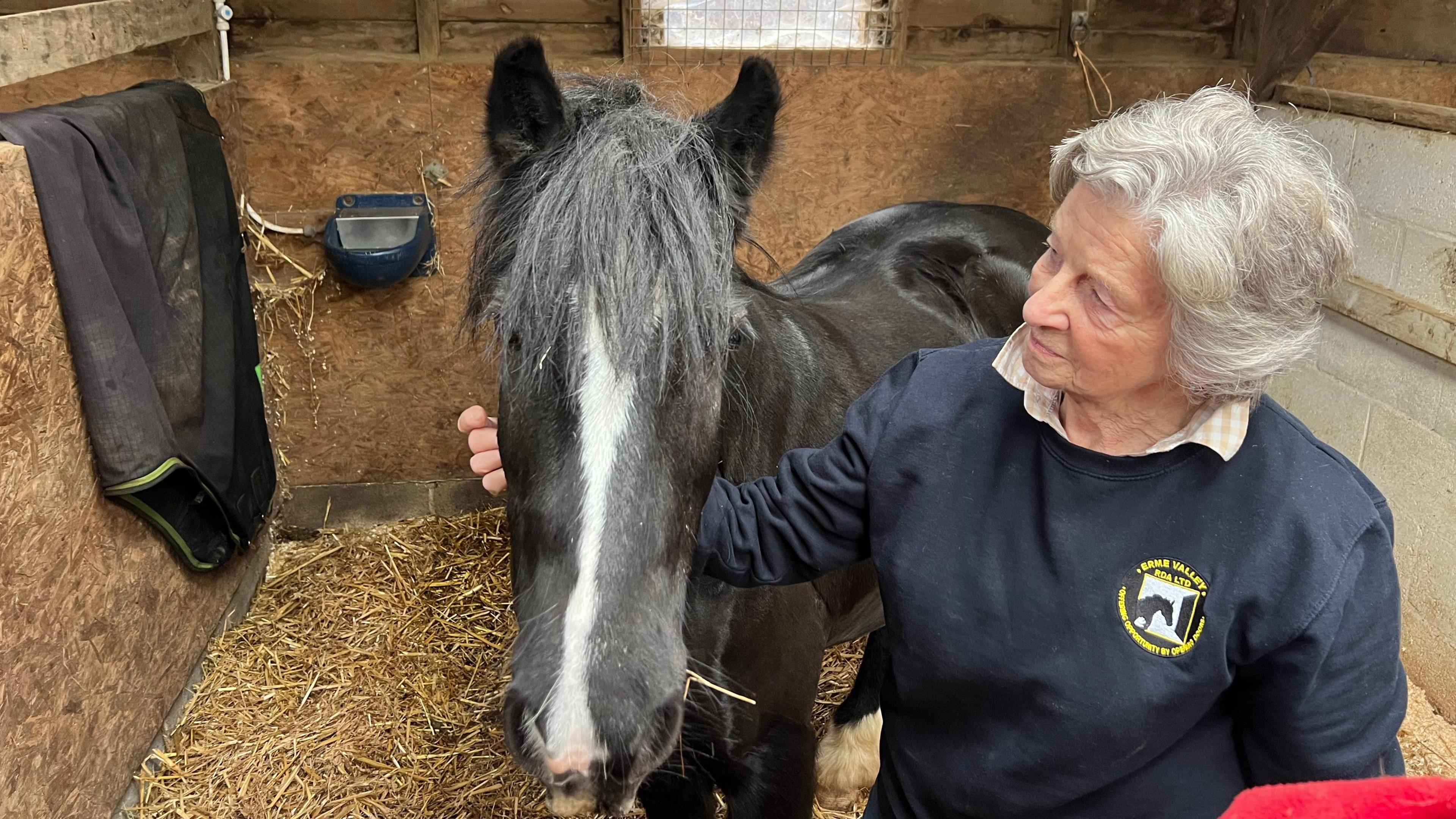 Sue Veale stroking a black and white horse in a stable. The horse has long black hair on its face and is on Sue's right. She is wearing an Erme Valley Riding for the Disabled jumper with the logo in the top right.