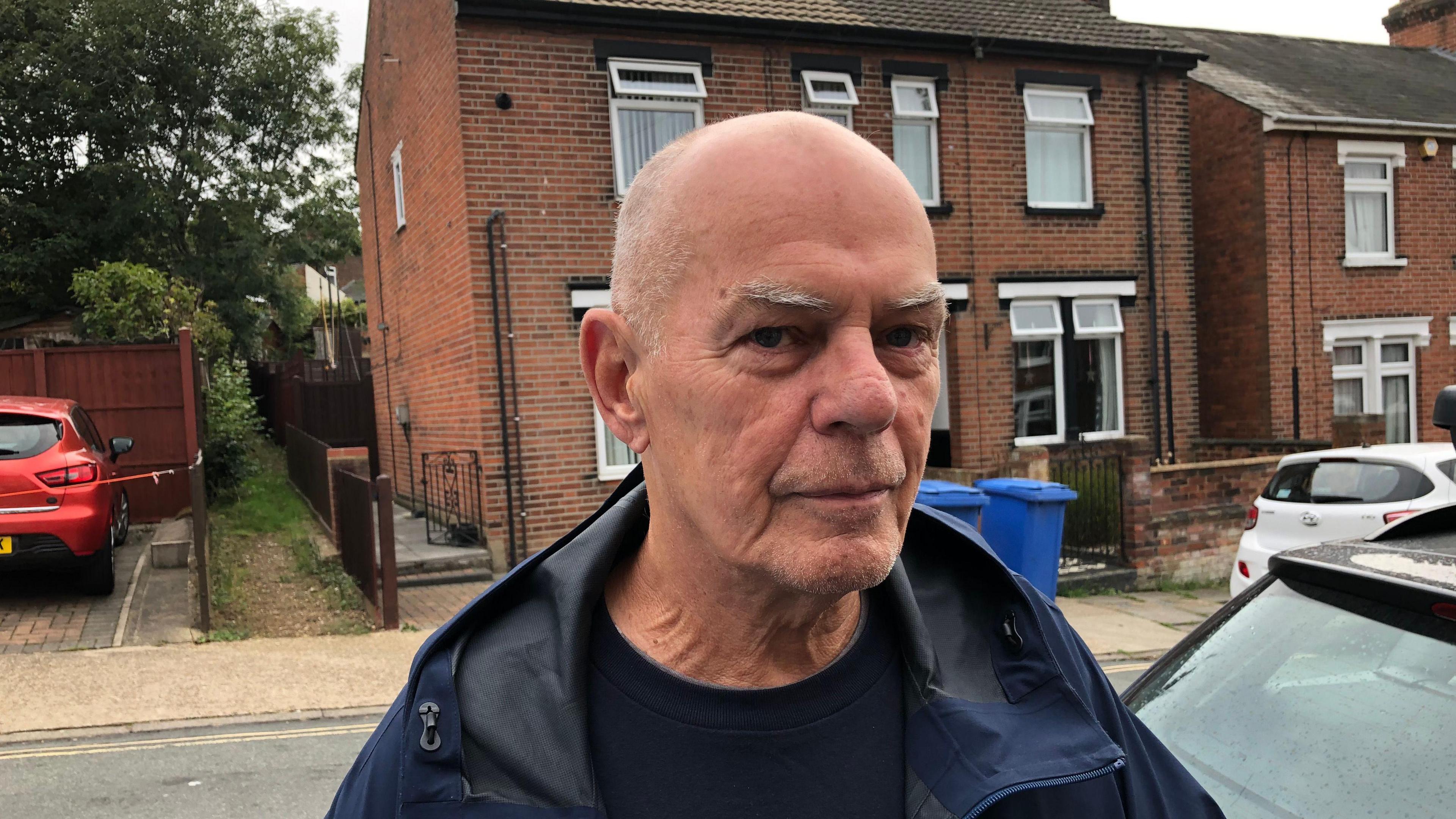 A picture of Nick Garnett on Devonshire Road in Ipswich. He is looking to the camera with an expressionless face. He is bald and is wearing a navy coat with a black t-shirt underneath. 