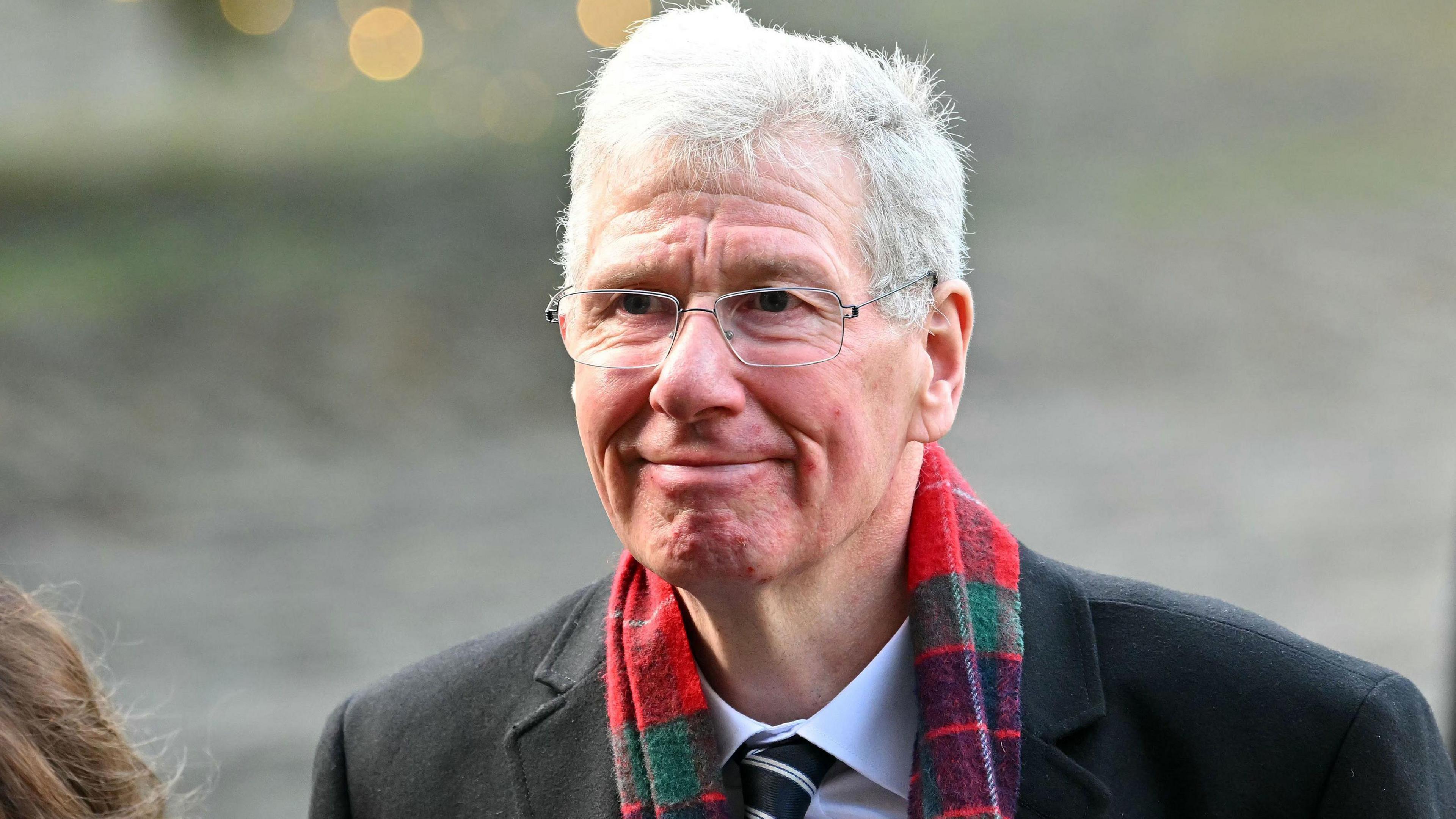 A smiling Kenny MacAskill, wearing a black coat, white shirt, black and white tie and a red tartan scarf, arriving at St Giles' cathedral in Edinburgh for the Alex Salmond memorial service. 