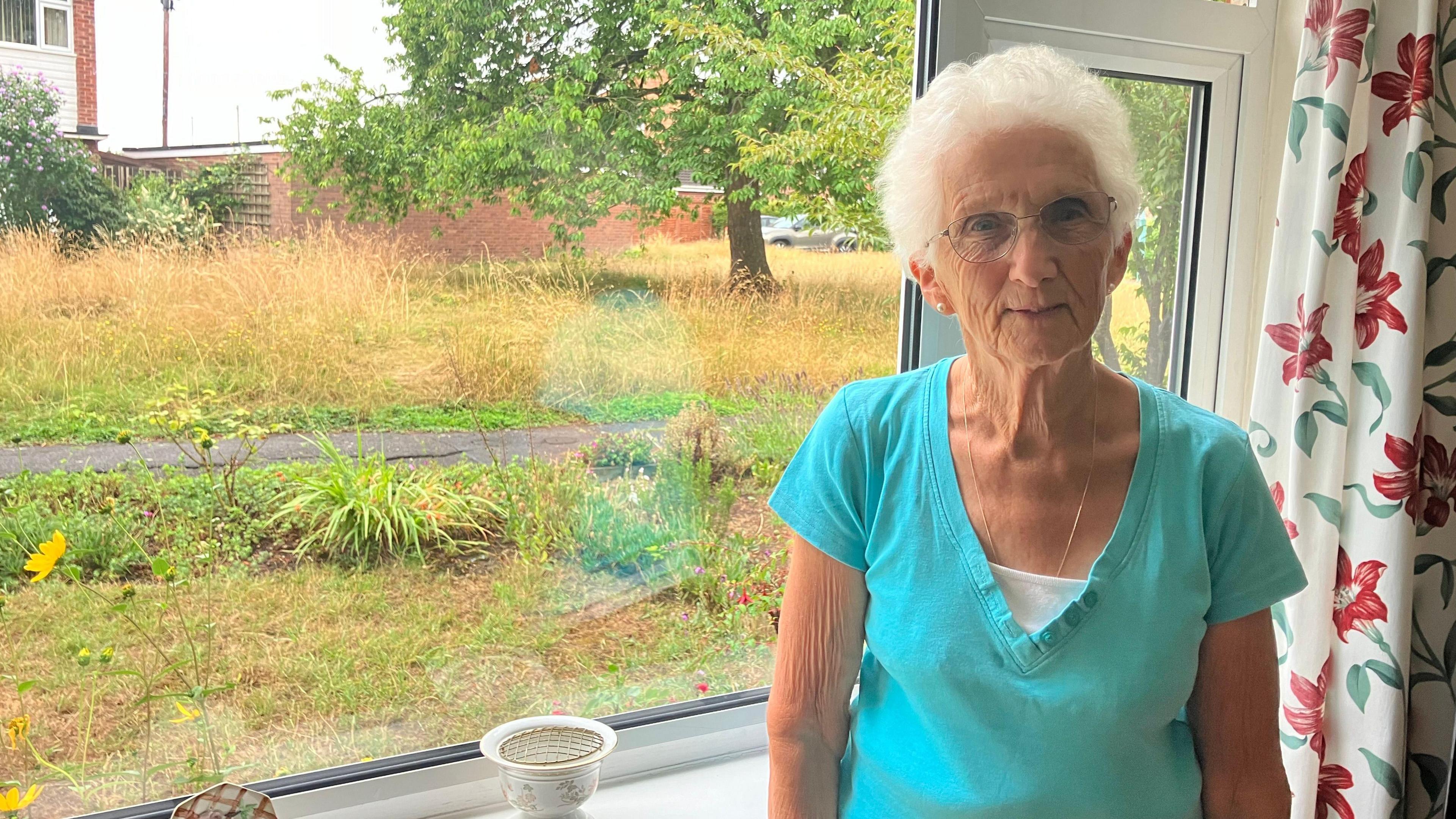 An elderly woman wearing a turquoise top stands in front of her living room window, which has white curtains with red flowers bordering it. Plants can be seen planted neatly around her front lawn. There is a tarmac path the other side of the garden, separating it from a large, overgrowing, brown area of grass, which has a tree growing on it. There is a house the other side of it.