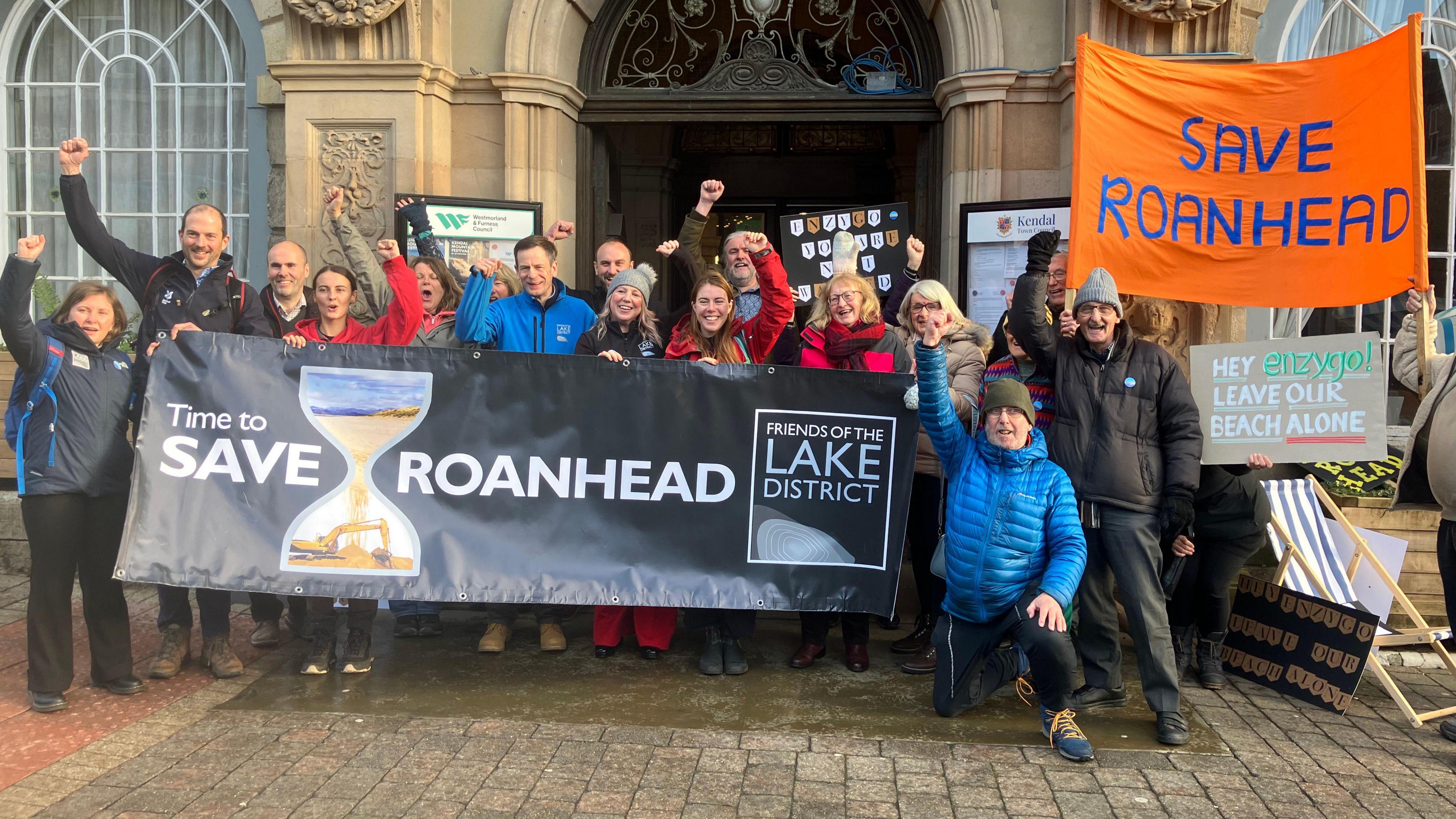 A group of men and women stand outside a council building, cheering, holding banners that read 'Save Roanhead'. 