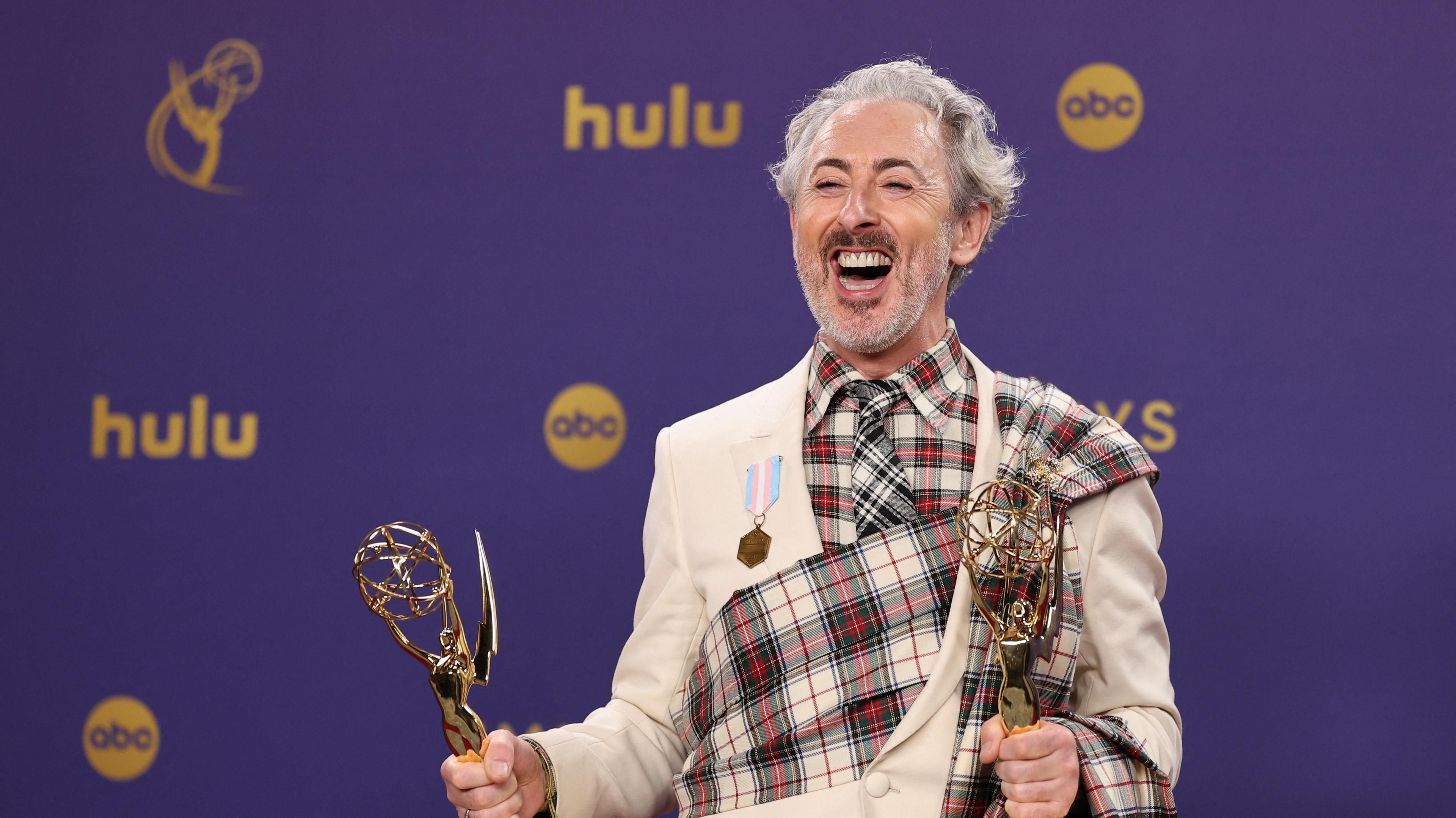 Alan Cumming, wearing a tartan wrap around a beige suit, celebrates winning the Outstanding Reality Competition Program for "The Traitors",