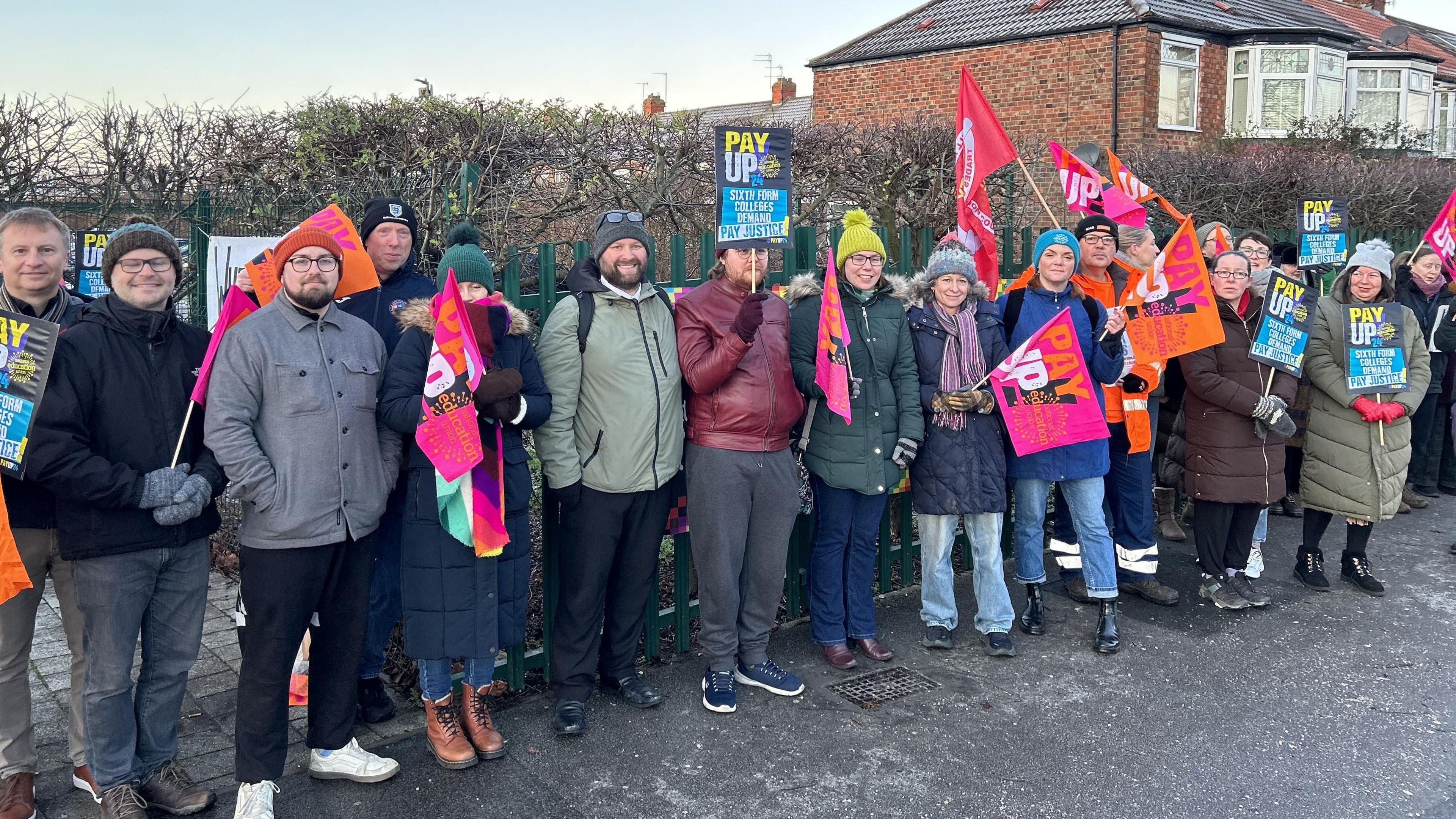 A group of 16 men and women wearing winter clothes stand on a picket line waving pink and orange-coloured union flags