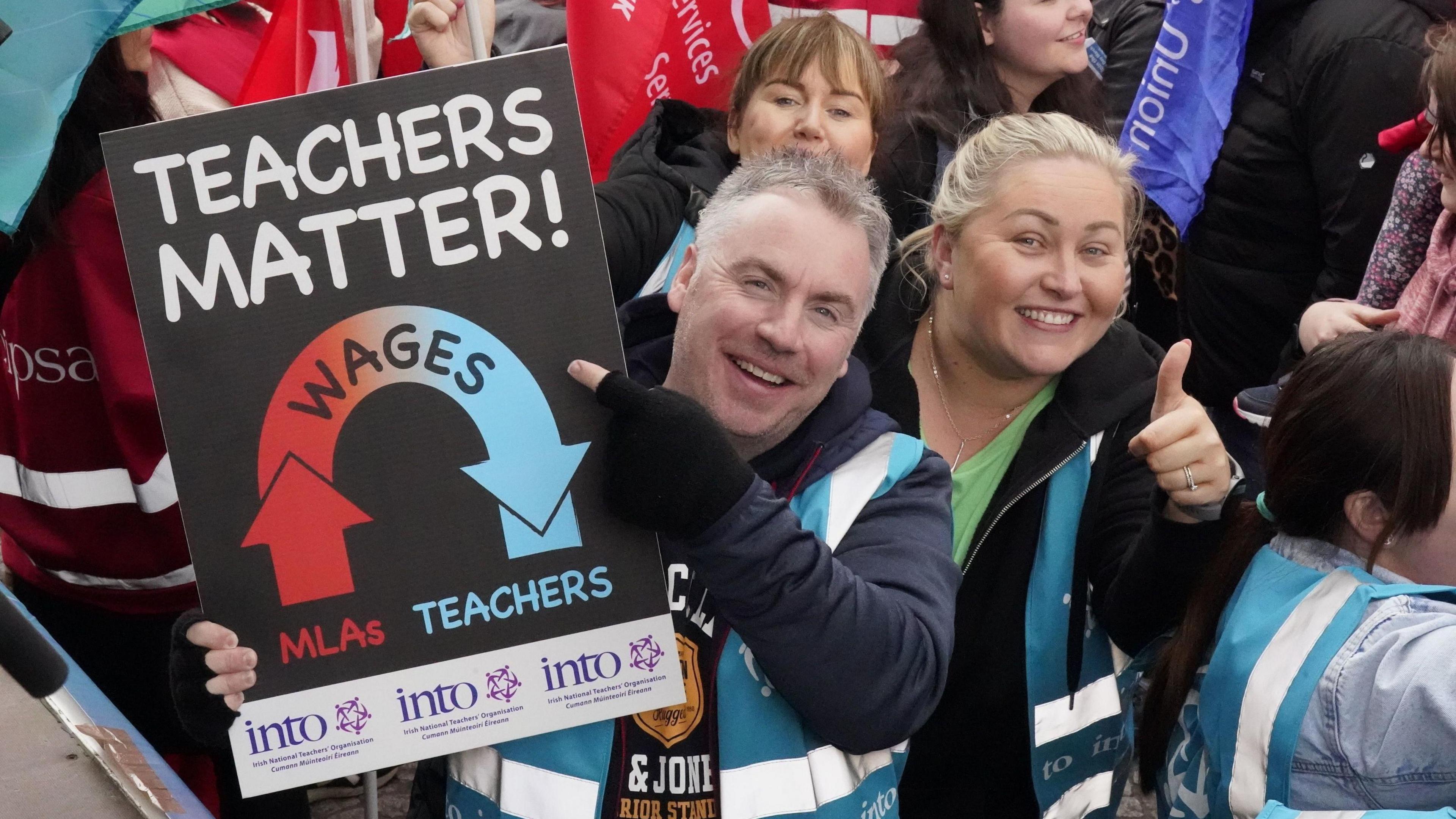 A man is smiling at the camera while wearing a blue hi-vis vest and holding up a sign saying 'Teachers Matter' along with a graphic showing MLA wages being trickled down to teachers. On his left there is a woman smiling at the camera with her thumb up.