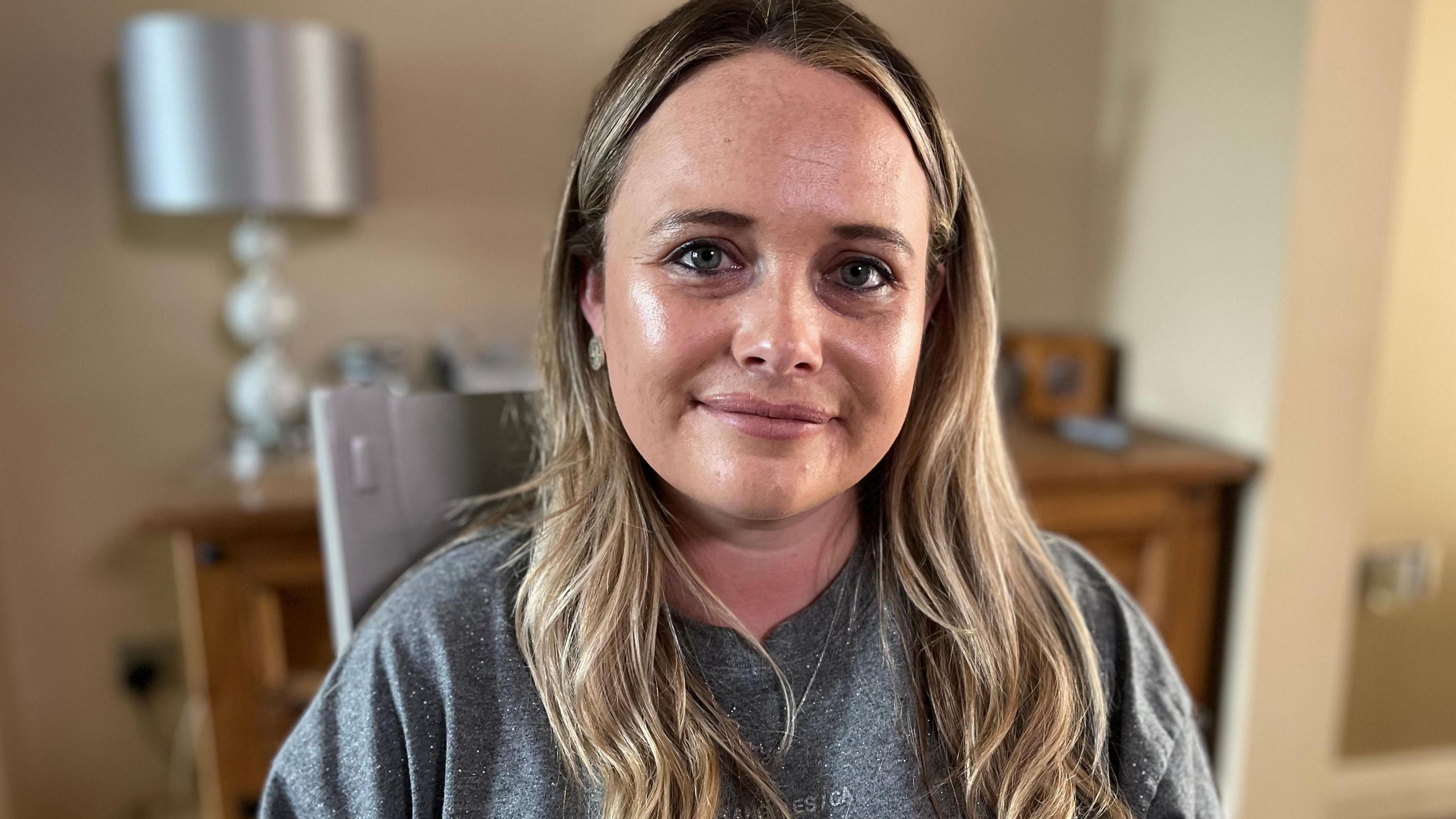 A woman with long very light brown to fair hair who is wearing a grey sweatshirt. She is sitting in a living room and a chest of drawers can be seen behind her, with a lamp and other out of focus items. She is looking at the camera and appears about to smile. 