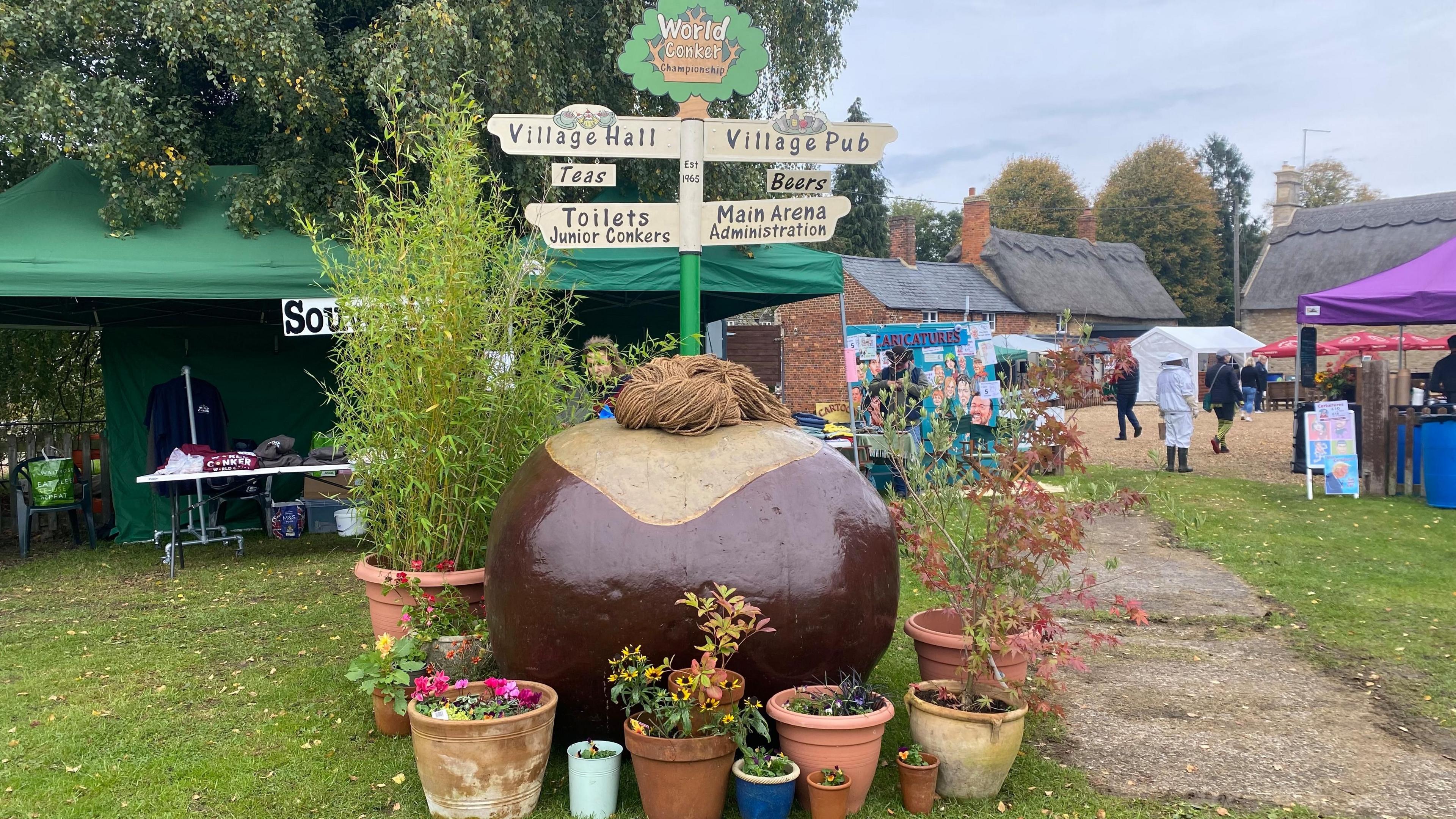 A view of the event. Stalls arer set up in a green common. There is a giant conker surrounded by plants with a sign coming from the top of it. The sign has directions for the village hall, pub and toilets.