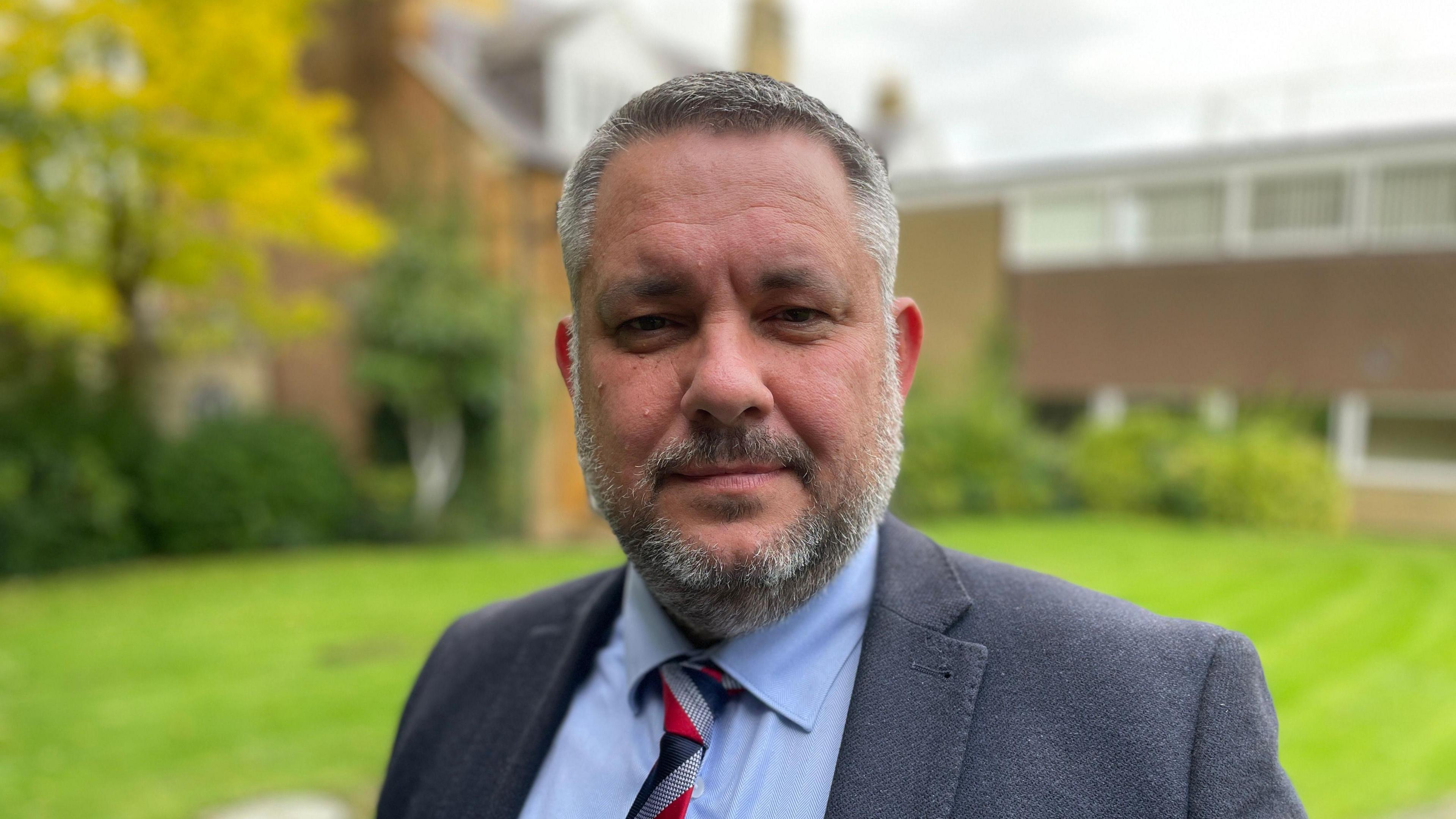 North Northamptonshire Council Leader Jason Smithers looks at the camera with a neutral expression. He is wearing a dark grey jacket with a light blue shirt and red, grey and blue striped tie. Behind him is a house and a lawn.