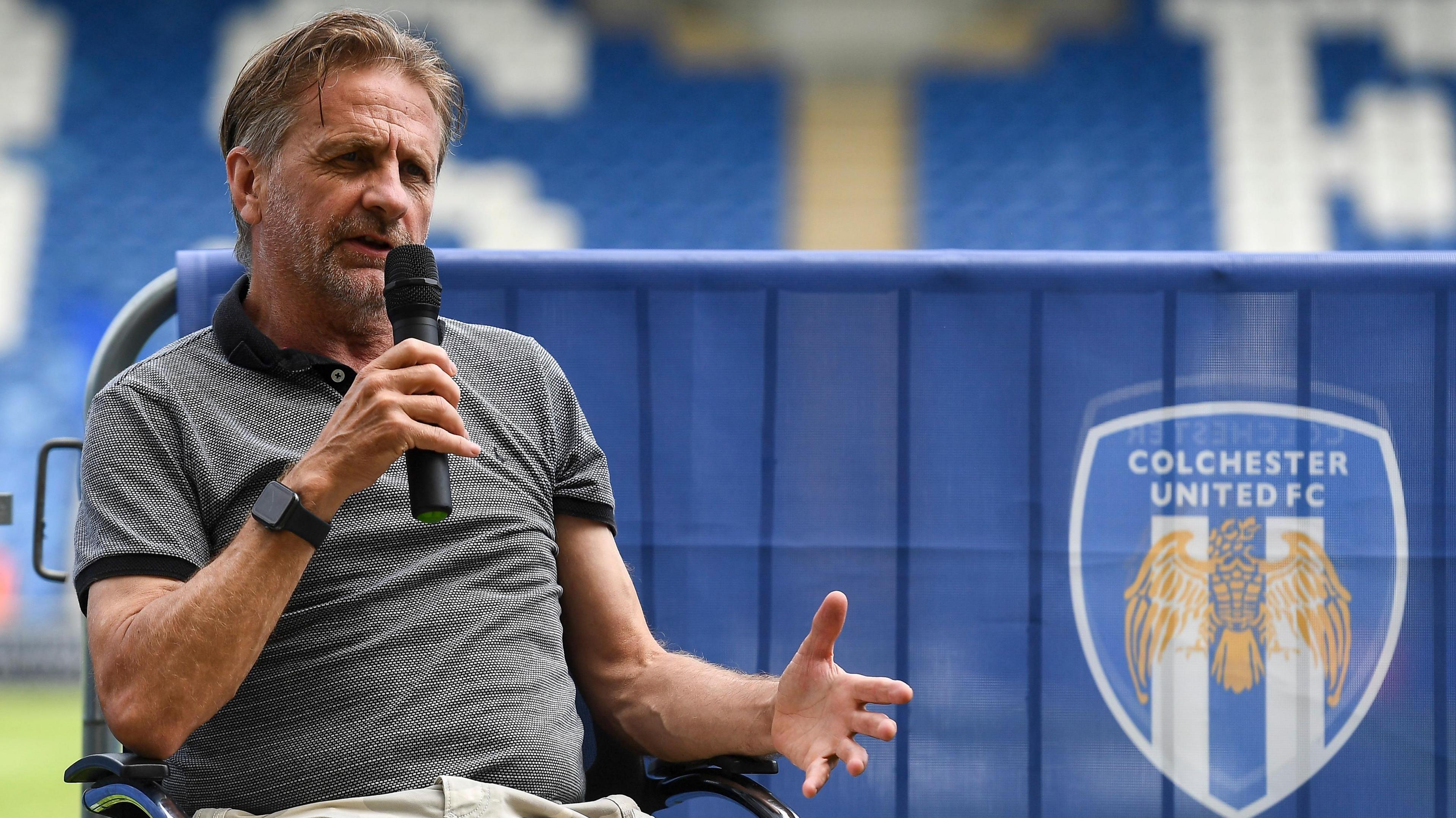 Colchester owner Robbie Cowling at the club's stadium, holding a microphone in his hand and speaking while seated