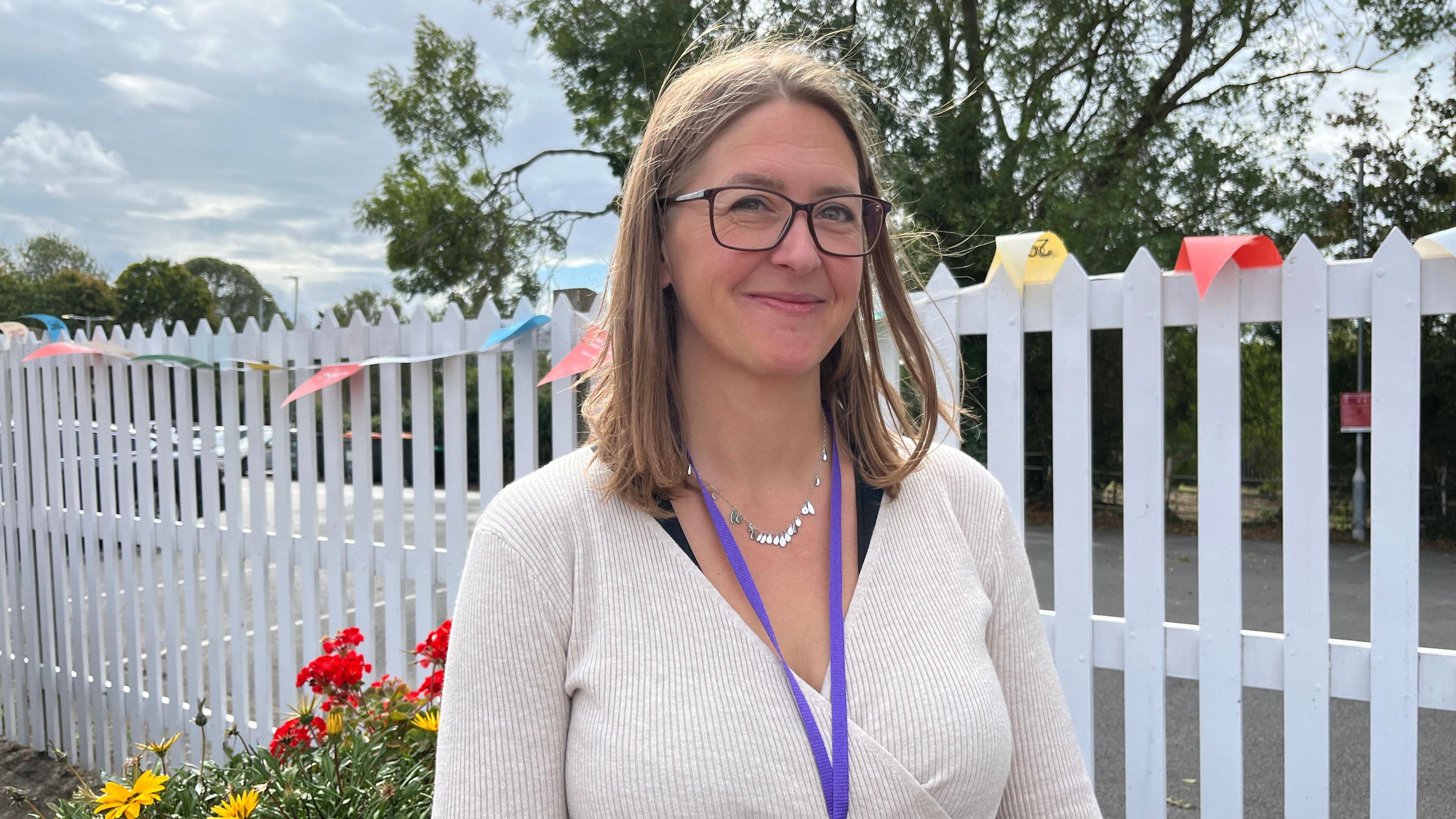 A woman standing on a platform at a railway station. She is wearing a cream pullover and has a purple work lanyard and a silver necklace around her neck. She has shoulder-length light brown hair. There are white railings behind her.