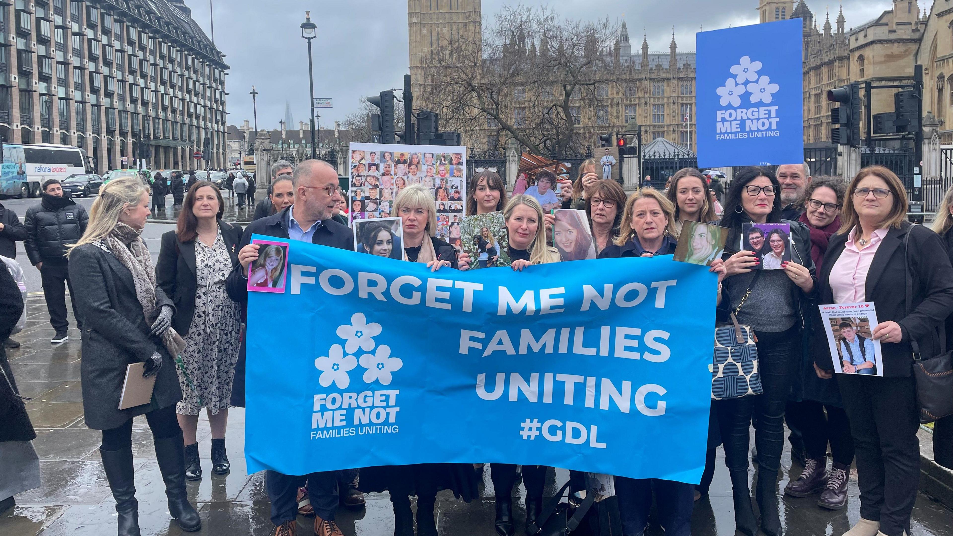 A gathering of men and women with a large blue flag in front of the Houses of Parliament. The flag reads forget me not families uniting.