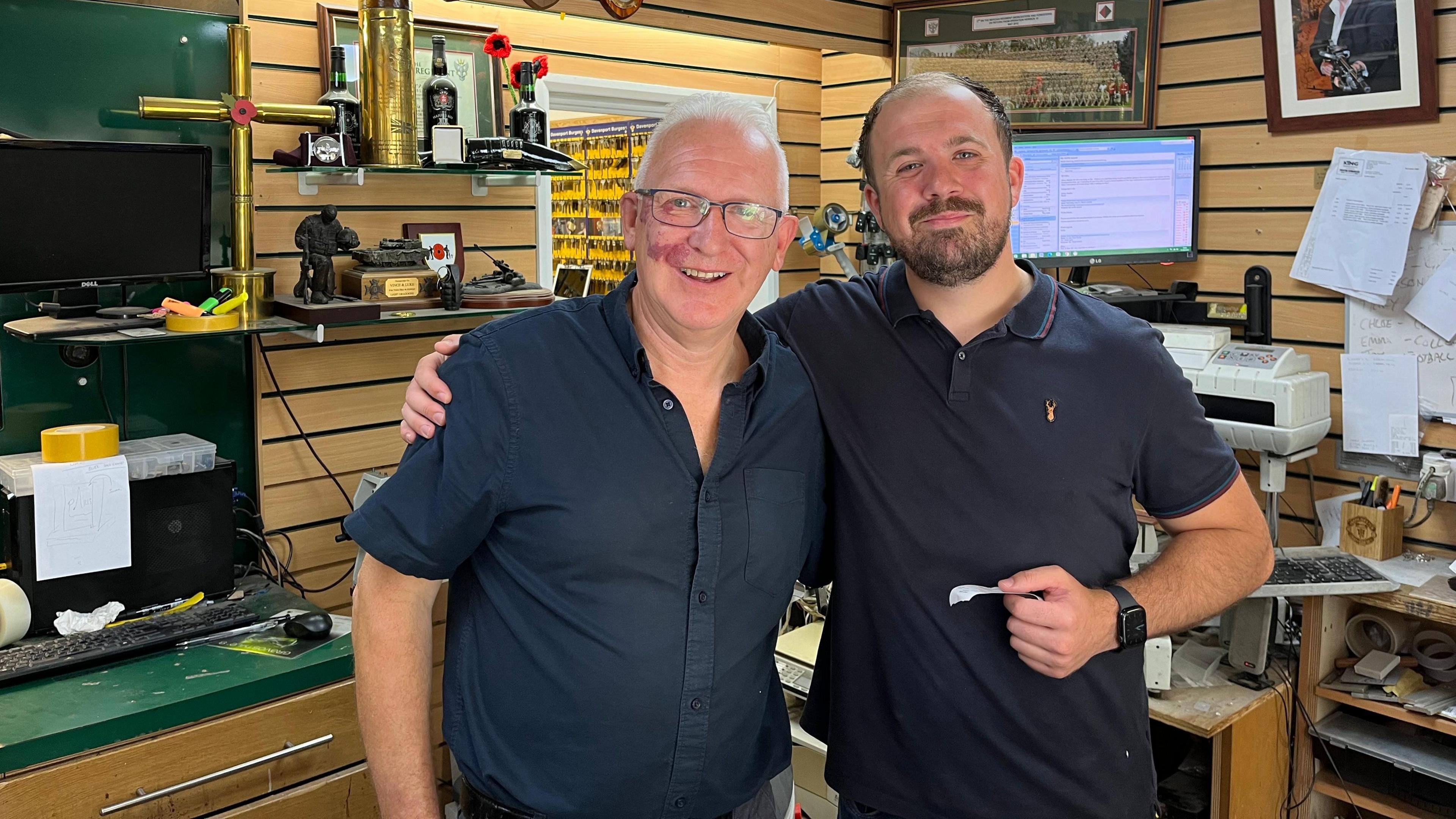 Mr Aylis wearing a black short-sleeved shirt and standing with his arm around another man in his shop. In the background you can see engraving tools