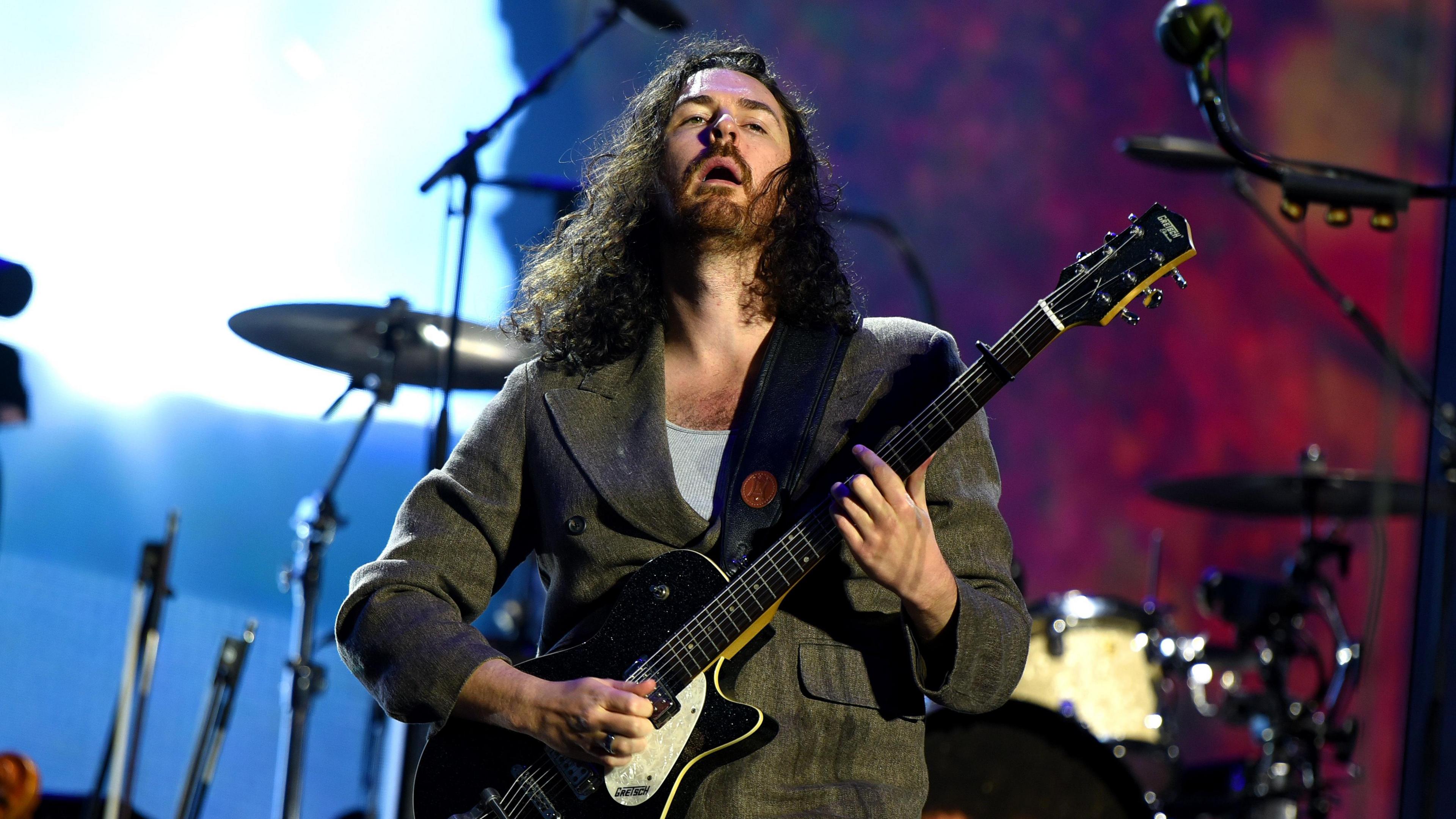 Hozier who has long curly hair and green jacket playing an electric guitar on stage. There is a set of drums behind him with a bright blue and dark red background.