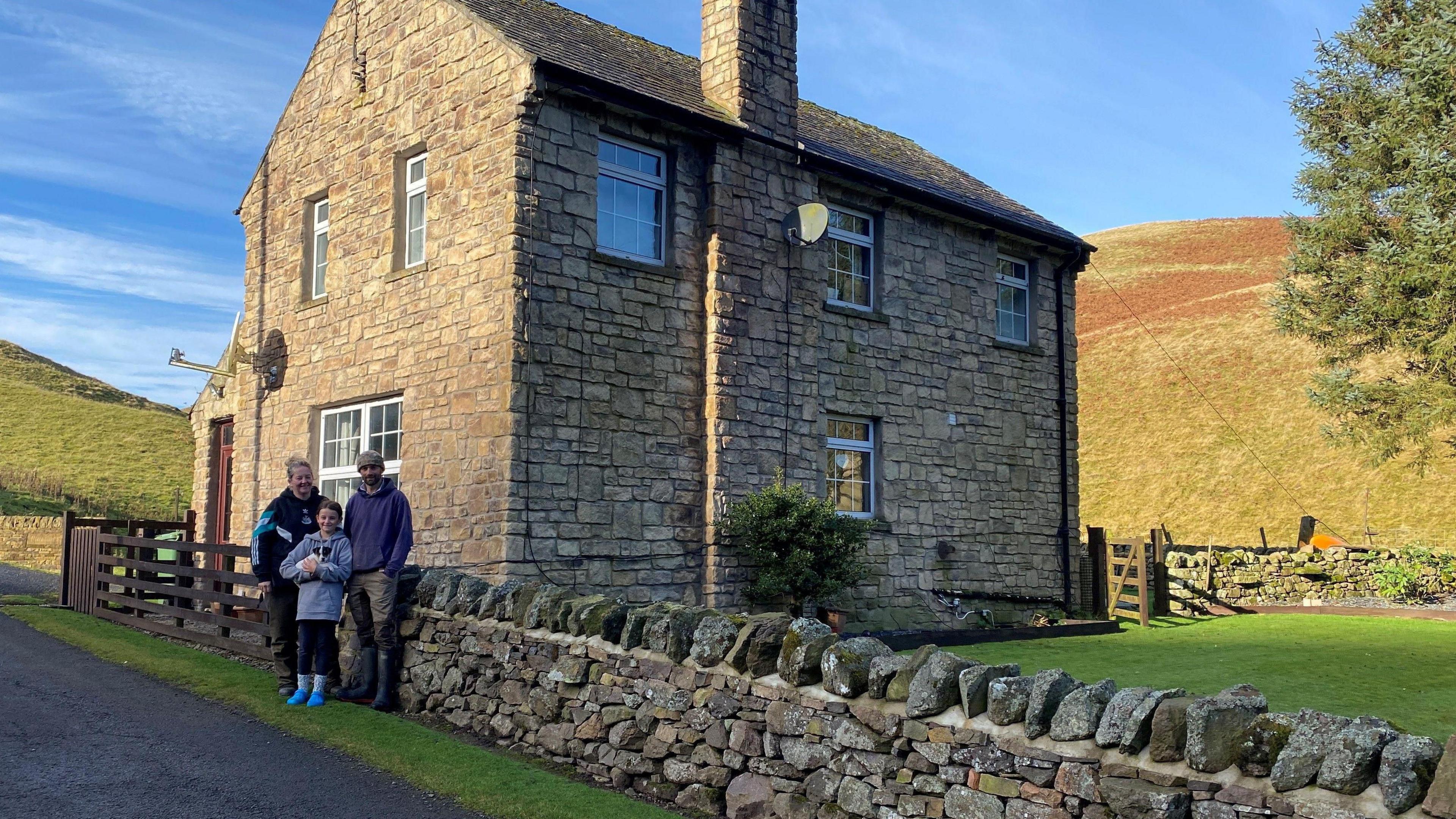 Harry and Megan Byatt are standing with a child outside a modern, two-storey stone farmhouse next to a single track road. Behind them the Cheviot hills and the house are bathed in late evening sunshine