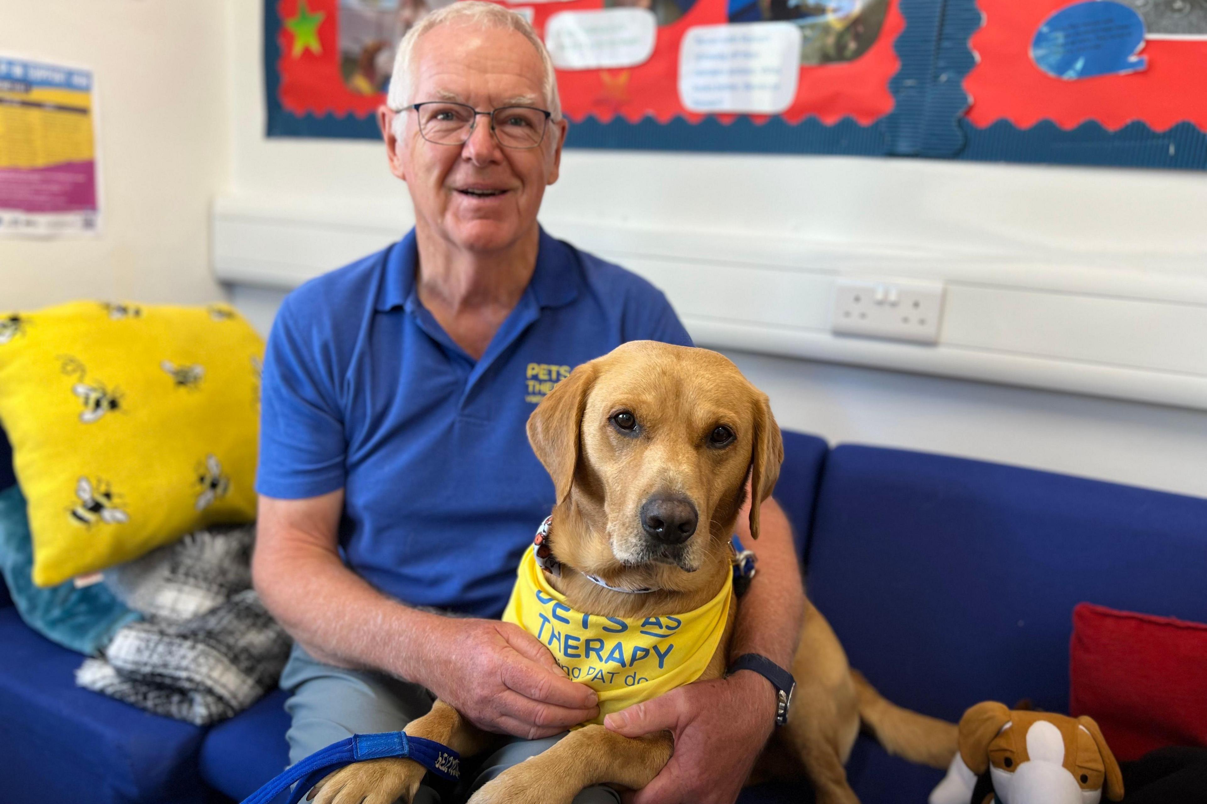A man in a blue polo shirt sits on a sofa with a golden labrador retriever on his knee, sporting a Pets As Therapy neckerchief