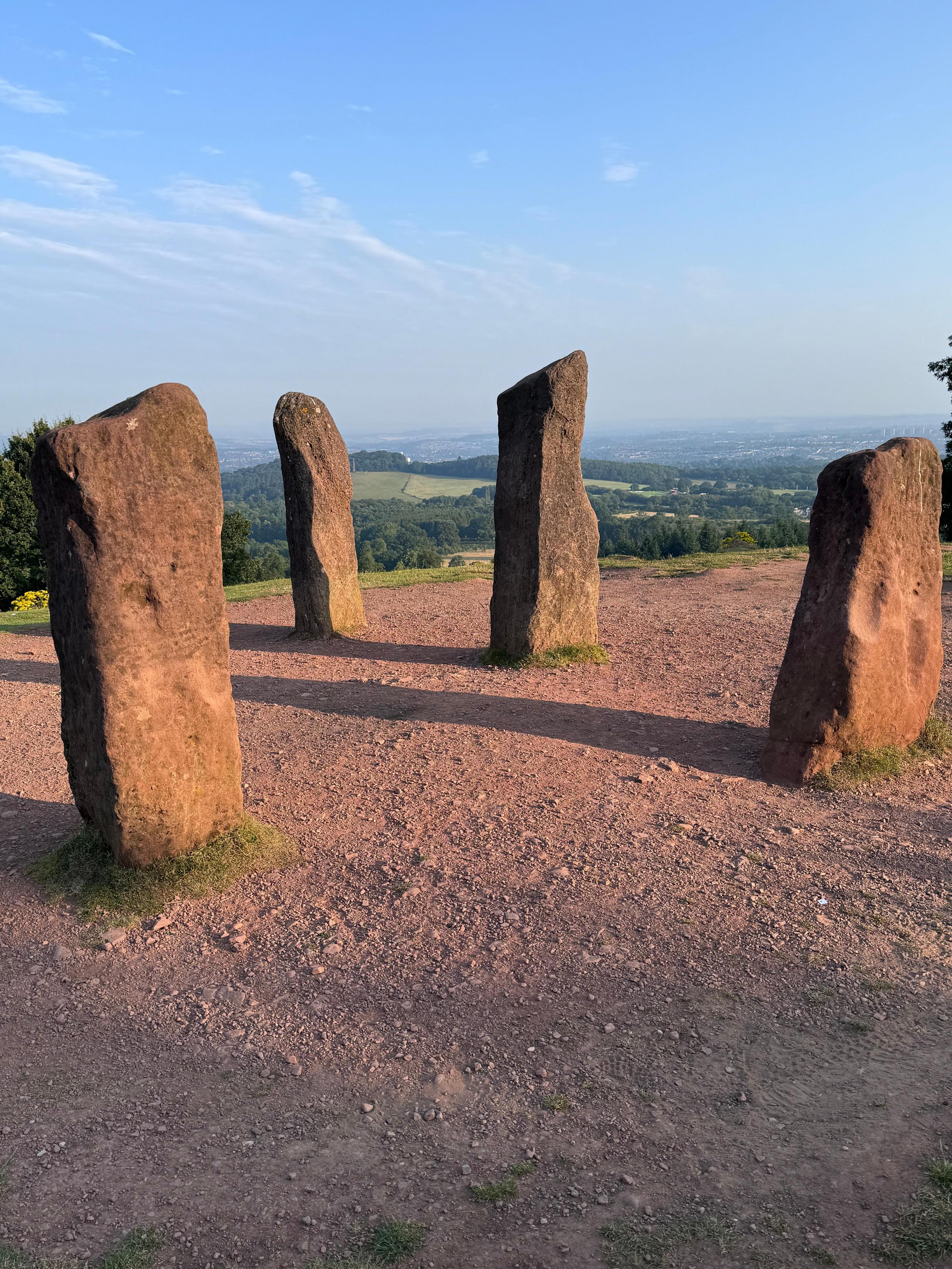 Four standing stones protrude from stony ground, with green hills and a blue sky in the distance  