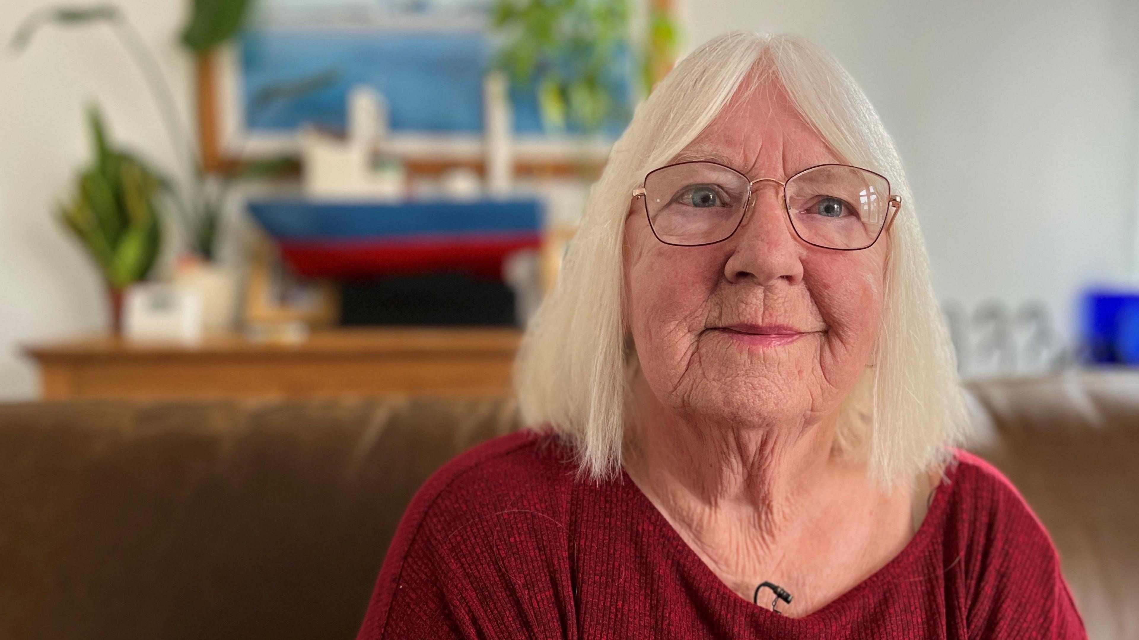 A woman sits in a smart-looking lounge. She has shoulder-length, white hair and wears glasses and a red top.