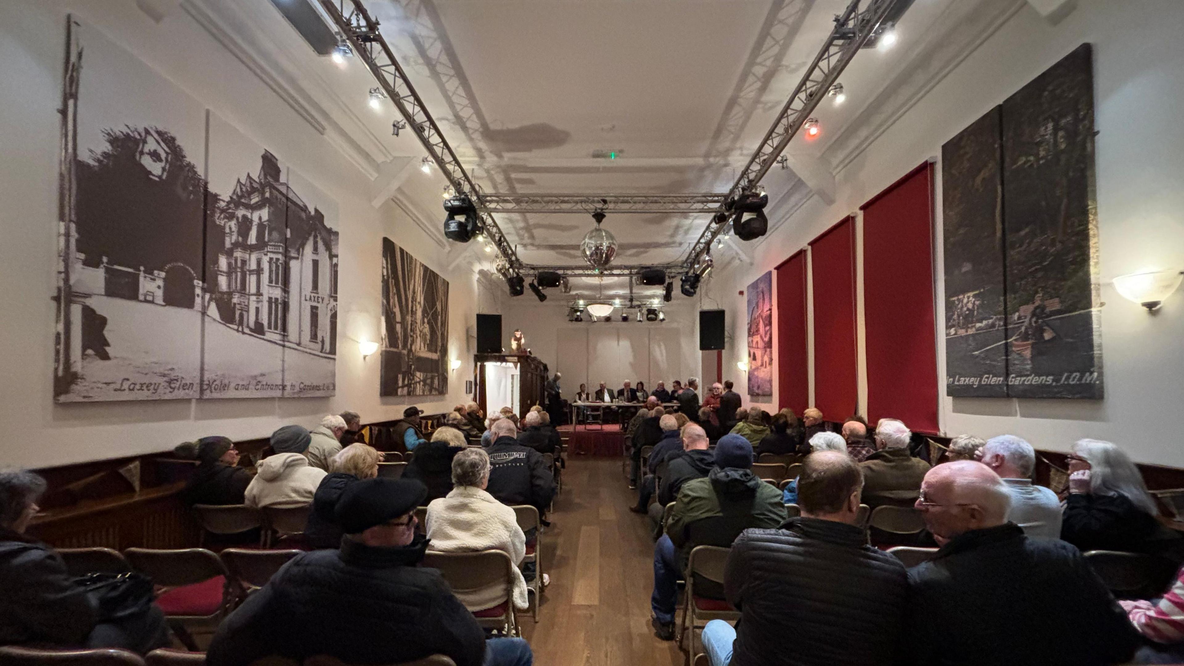 The meeting from the back, it is a long hall which is fairly full, there are a panel of people sat at a table at the front.