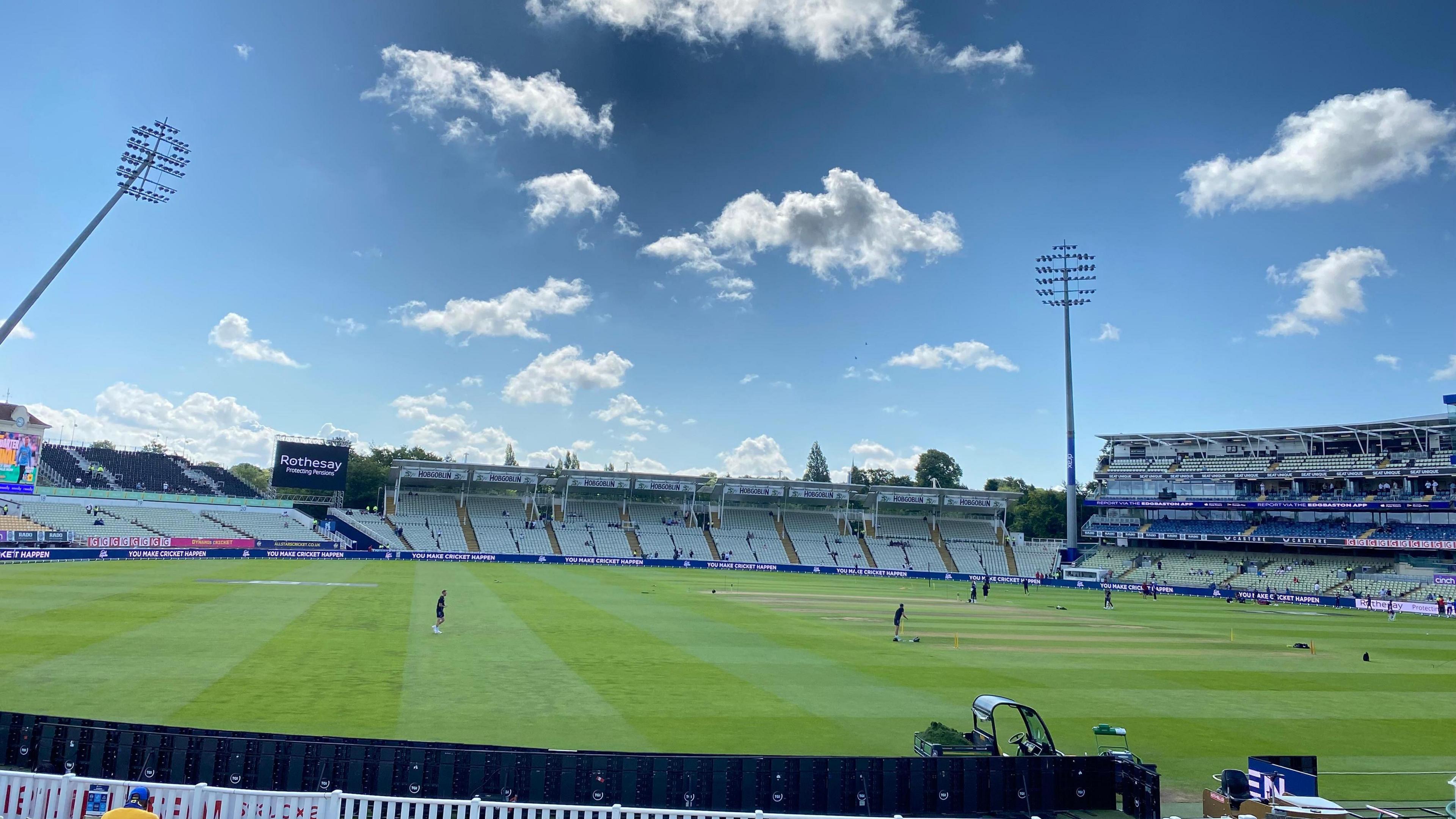 Wide shot of Edgbaston cricket ground, showing blue sky and white clouds, thinly-occupied stands and players practising on the pitch