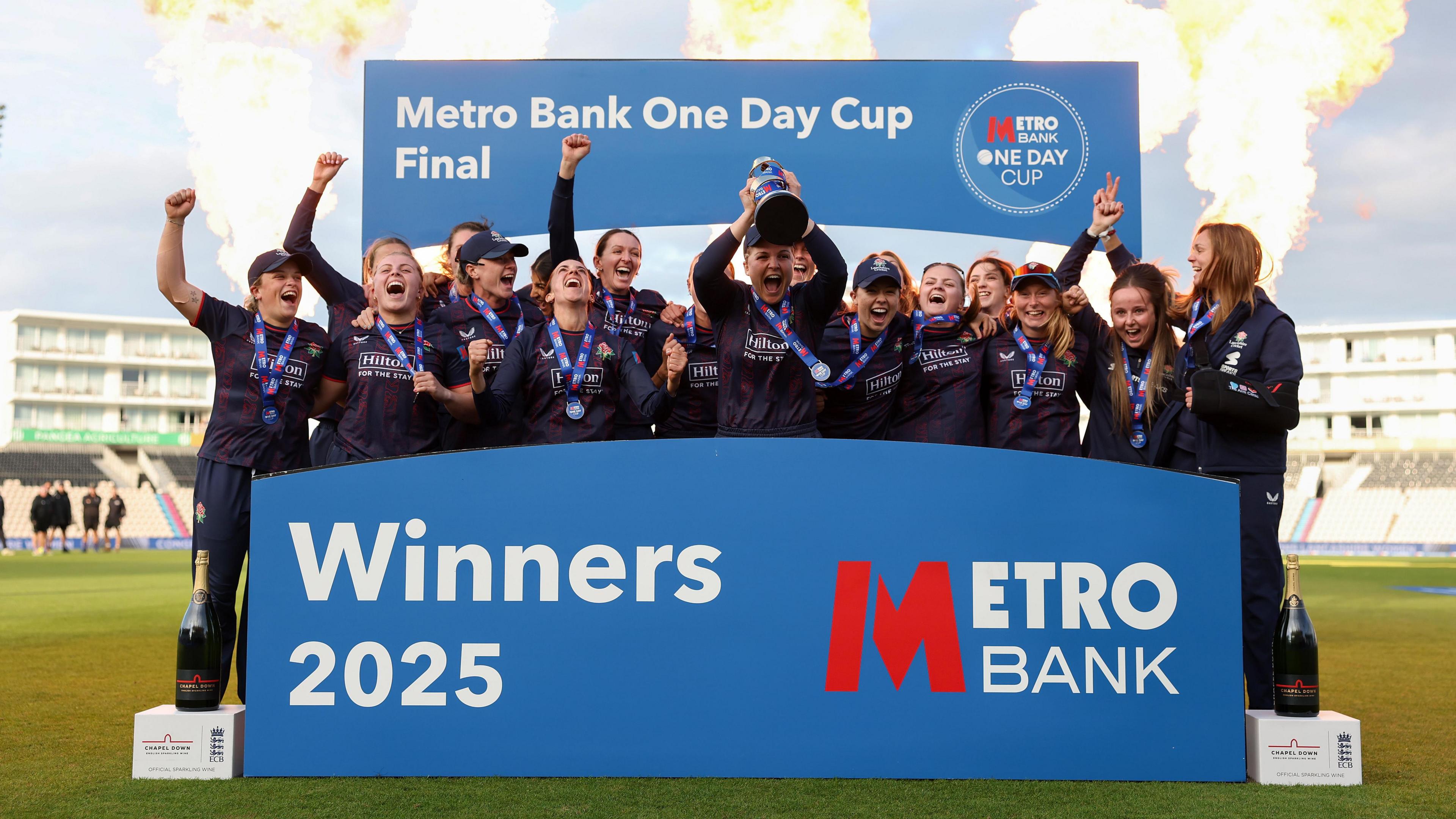 Lancashire Women lift the trophy and celebrate. There are flames in the background and the team stand behind a blue Metro Bank Advertisement board