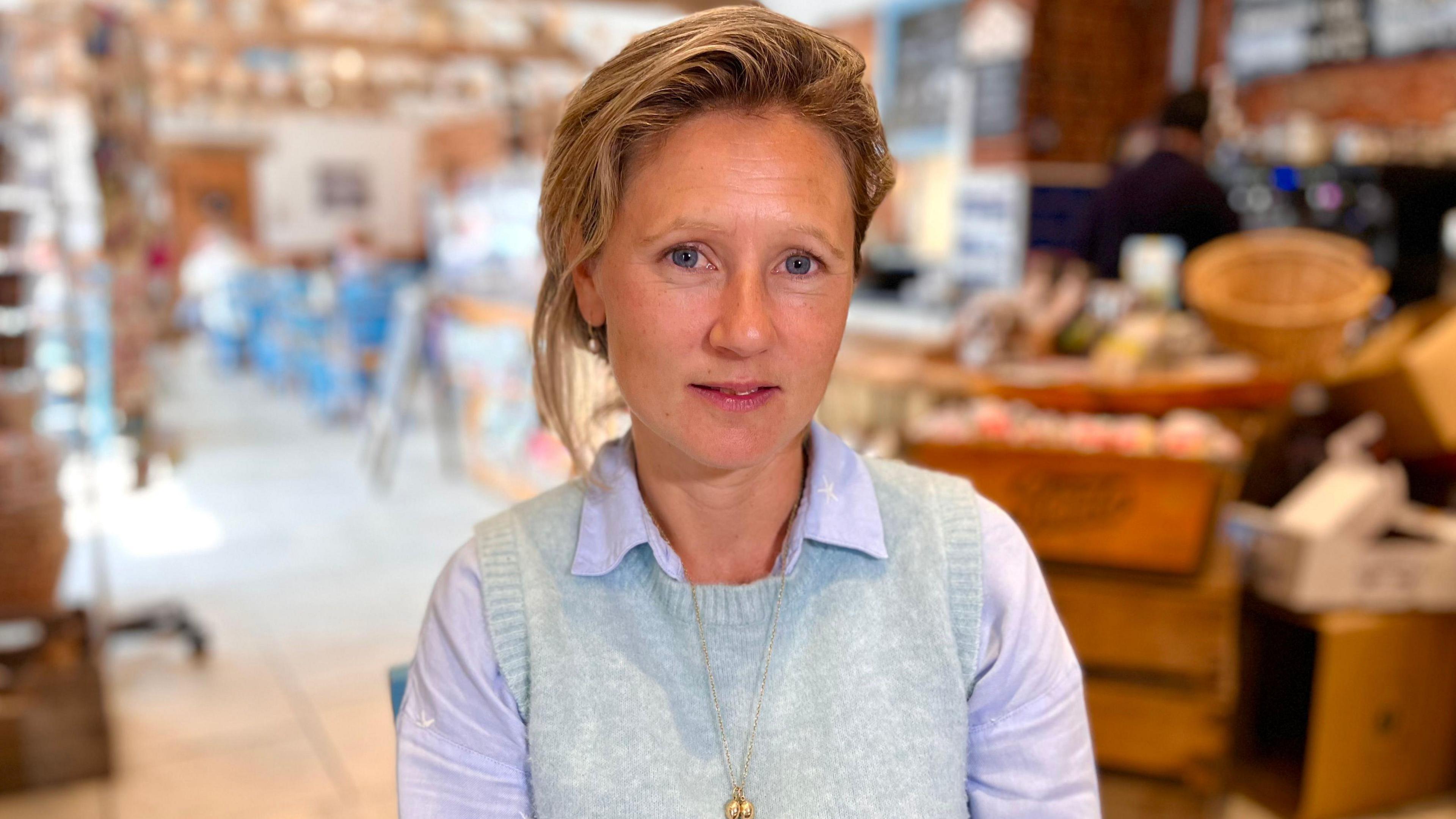 Charlotte Gurney wears a grey tunic jumper over a purple collared shirt as she sits amongst produce in the farm shop