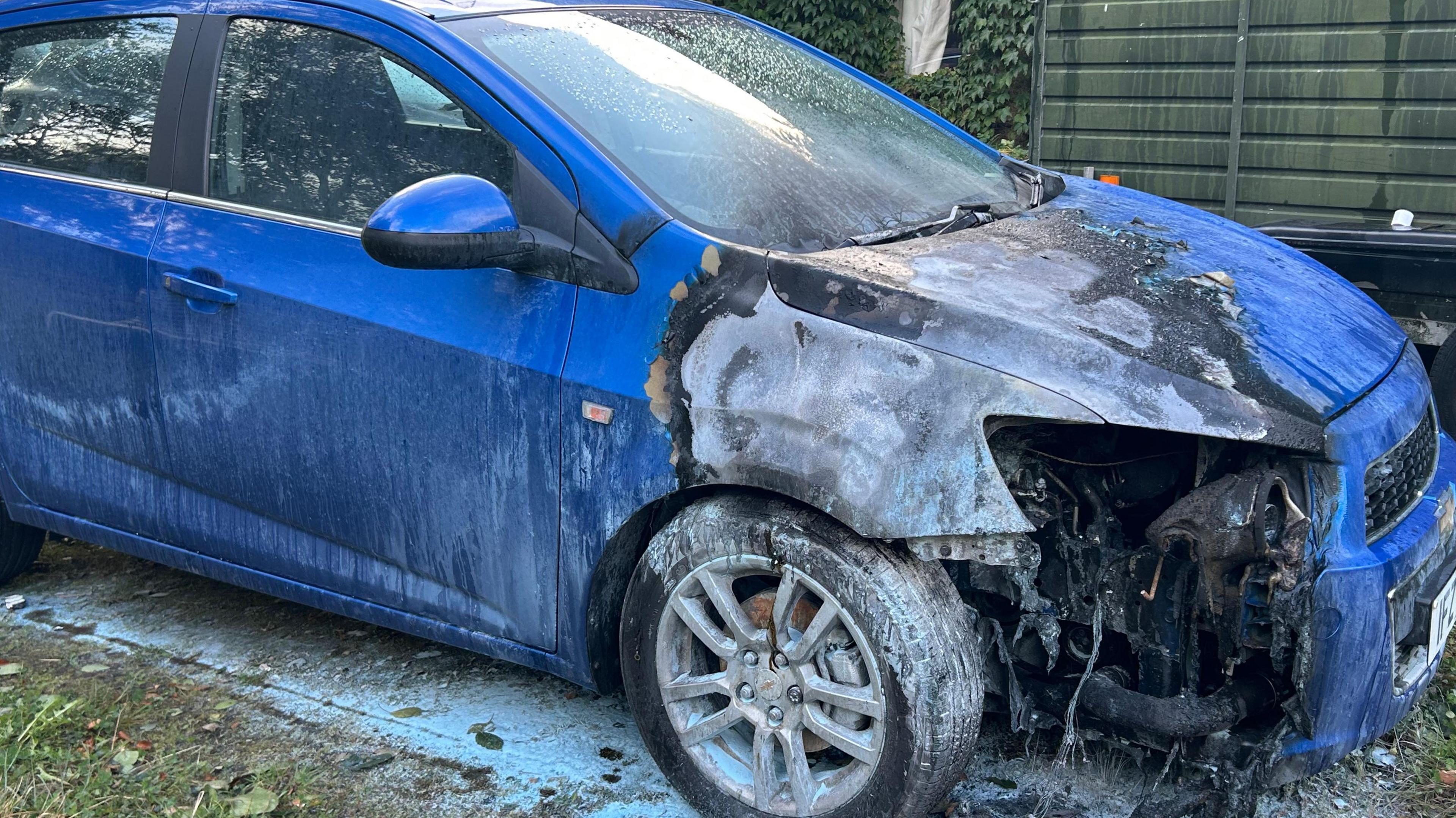 A blue car has the front left bumper burned to appear grey. The wheel is covered in white ash.