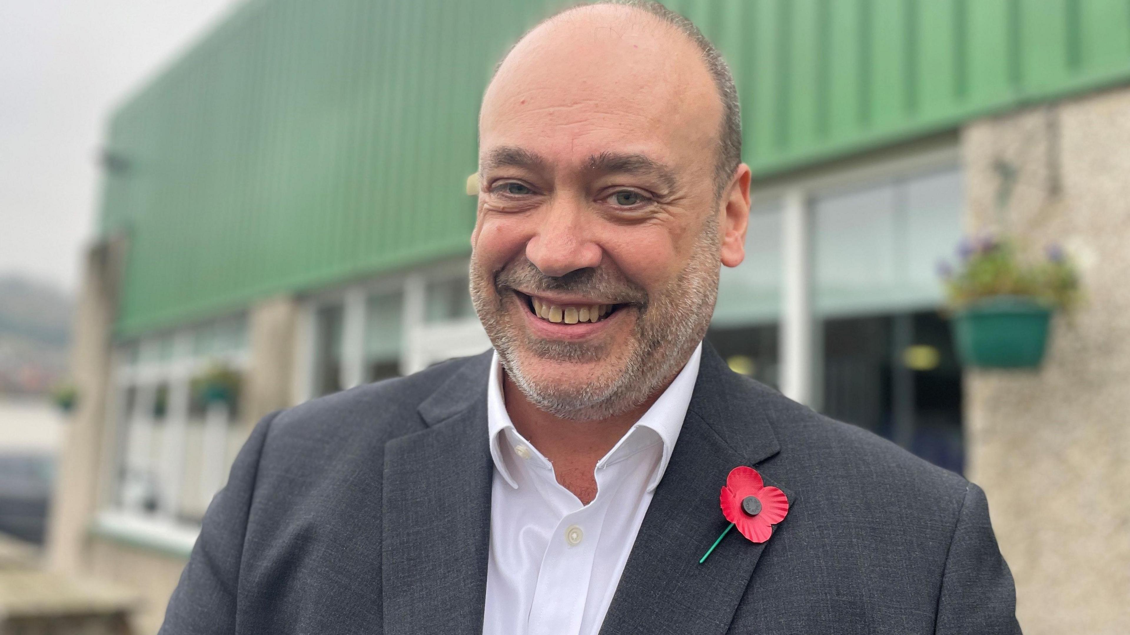 A smiling man with a bald head and greyish stubble in suit jacket and white shirt unbuttoned at the neck wearing a poppy