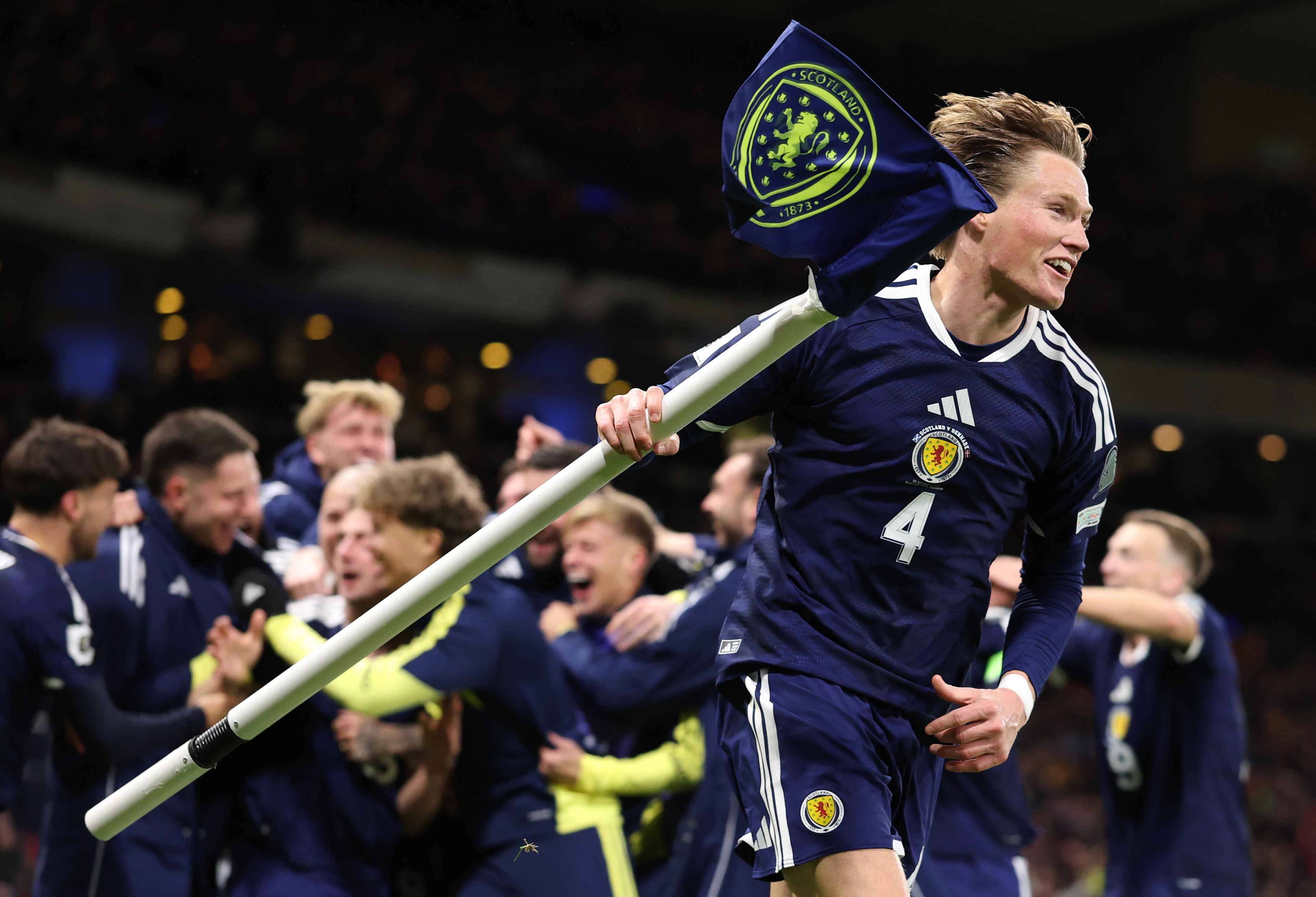 Football player in a dark blue Scotland kit holding a corner flag while team-mates celebrate in the background during a match.