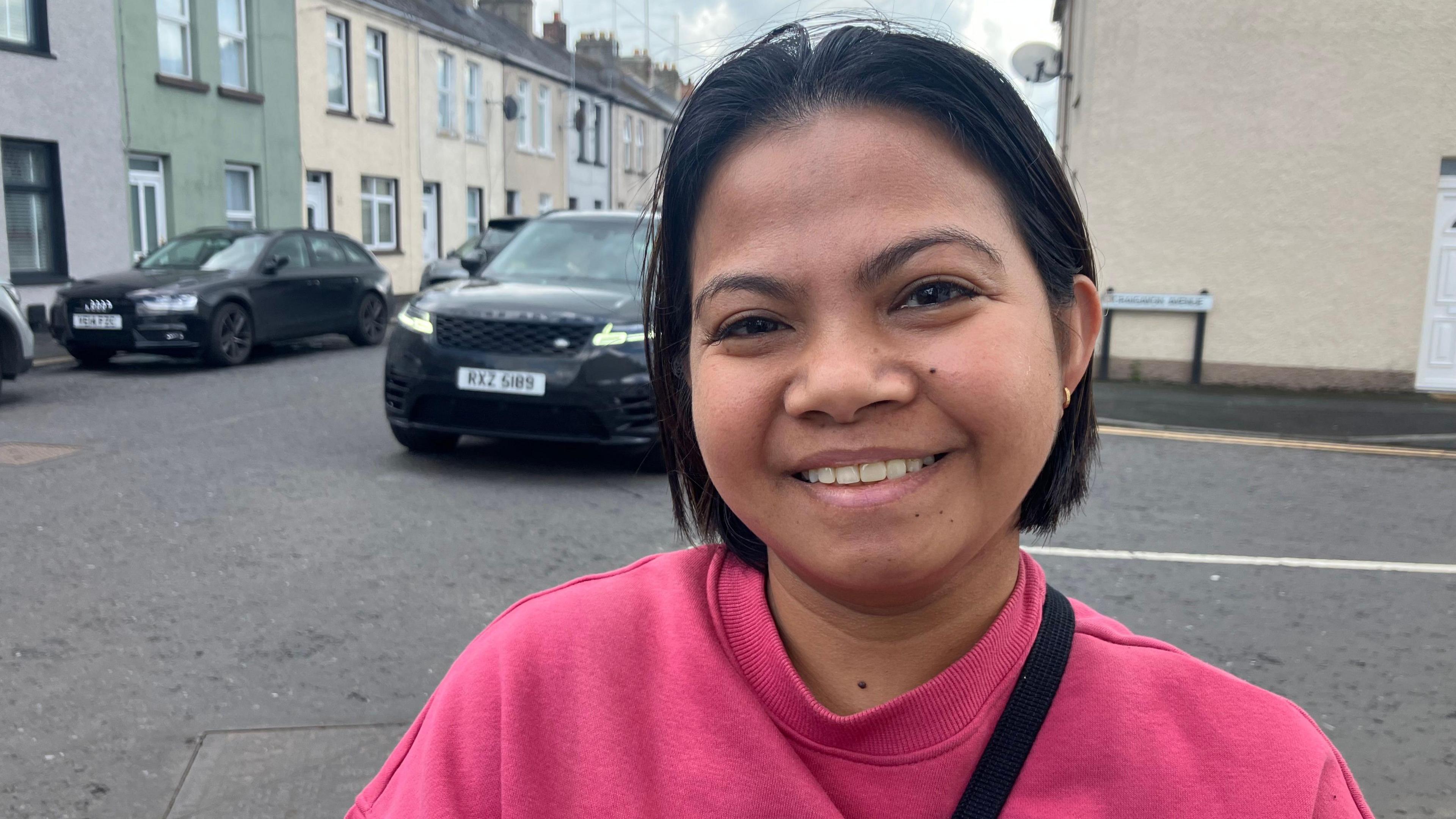 A woman smiles into the camera. She has short black hair and is wearing a pink jumper. She's standing in a street. Cars are parked behind her.