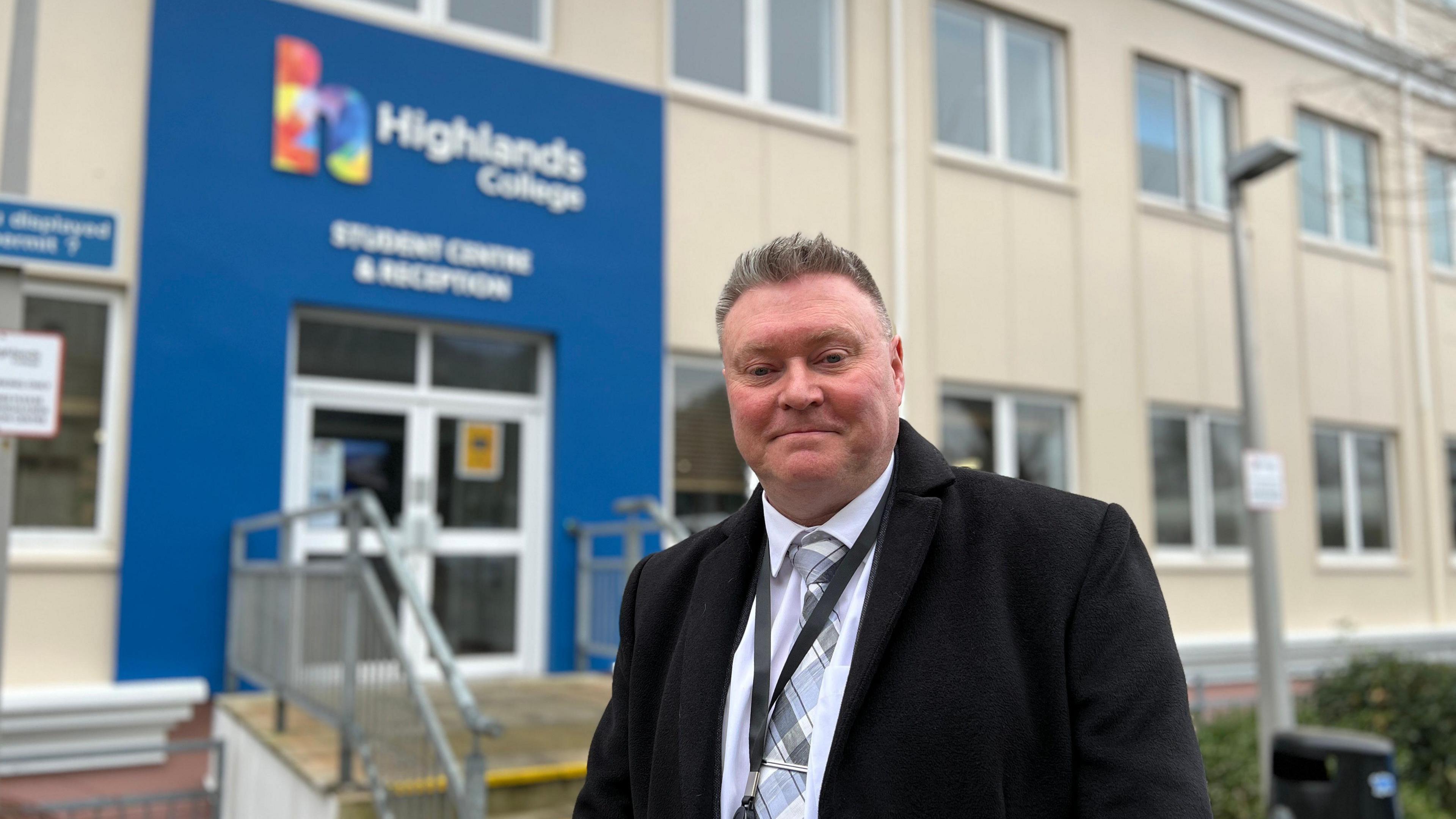 Deputy Rob Ward, a man with short grey hair. He is wearing a dark overcoat, a white shirt and a tie with varying shades of grey, white and light blue lines. He has a black lanyard string around his neck. He is standing outside a building with lots of windows, each with a white frame. Behind him is a set of steps with metal railings, and a wall painted royal blue. The sign above it says 'Highlands College', next to a large letter 'h' in multiple colours.