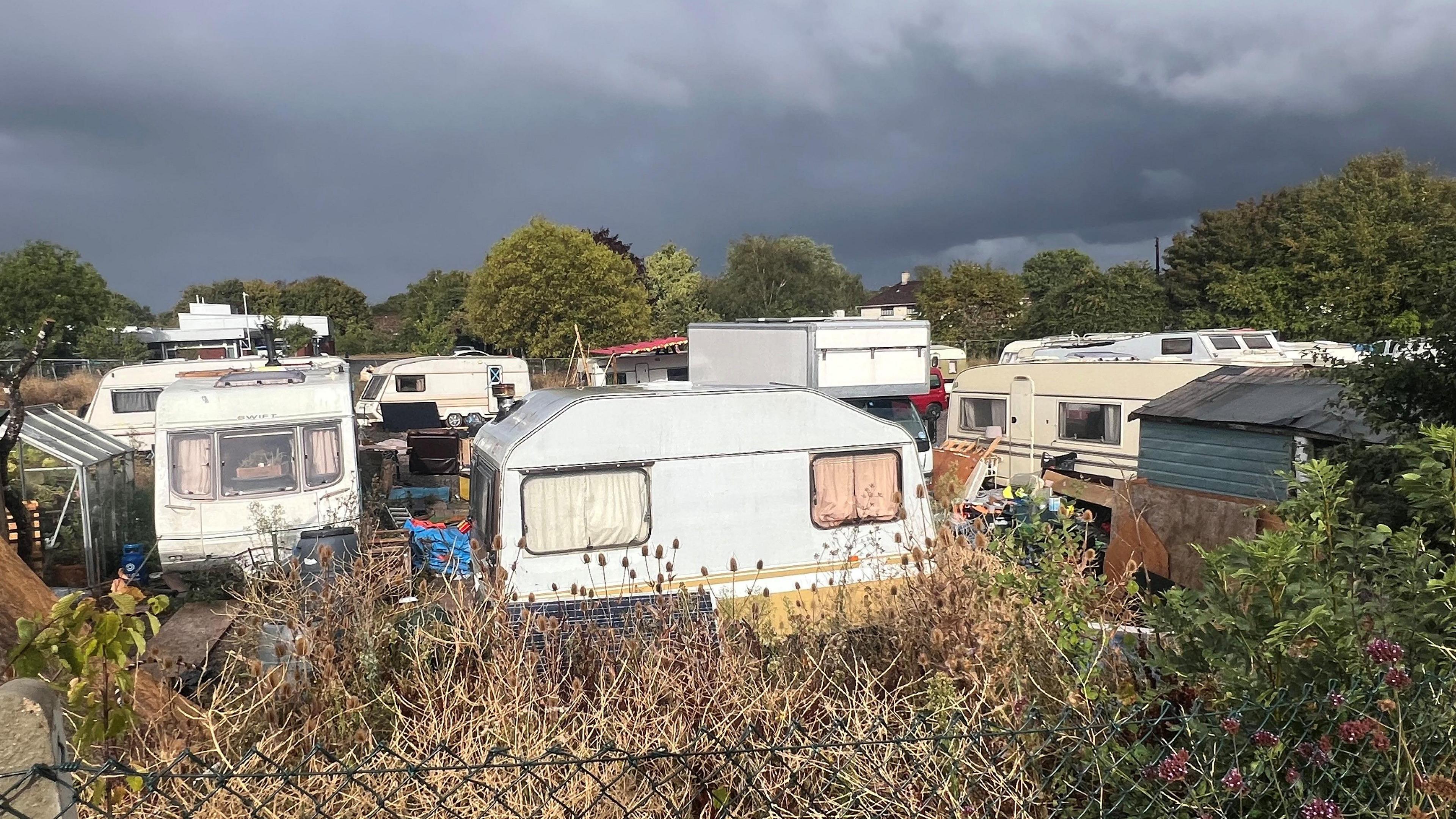A large group of vans, caravans and cars, alongside lots of miscellaneous items and possible litter parked up on a piece of land. The picture has been taken from behind a chain link fence.  