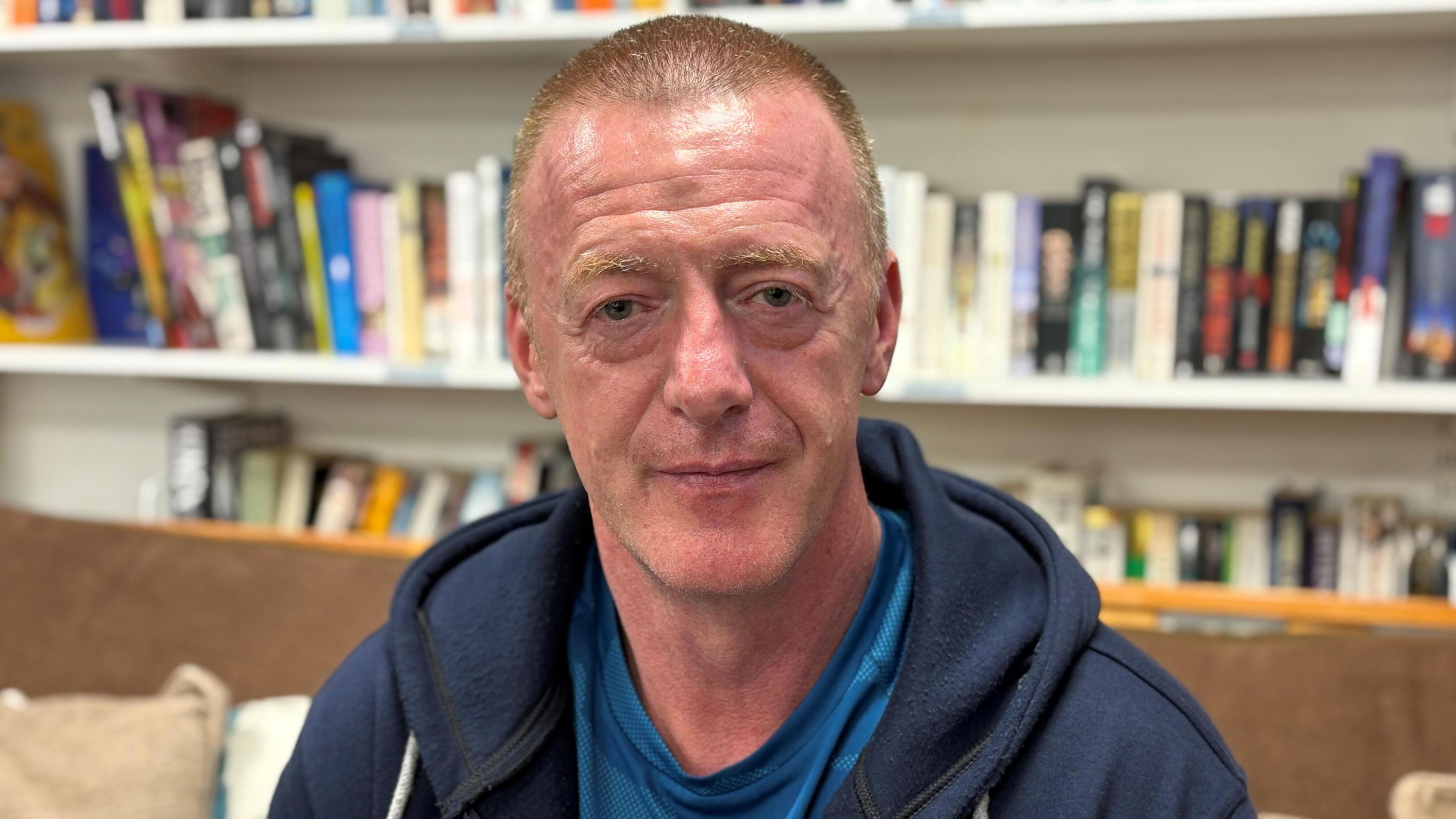 A man with short brown hair, wearing a blue hoody and blue t-shirt, sits on a brown sofa. There are bookshelves behind him, lined with books.