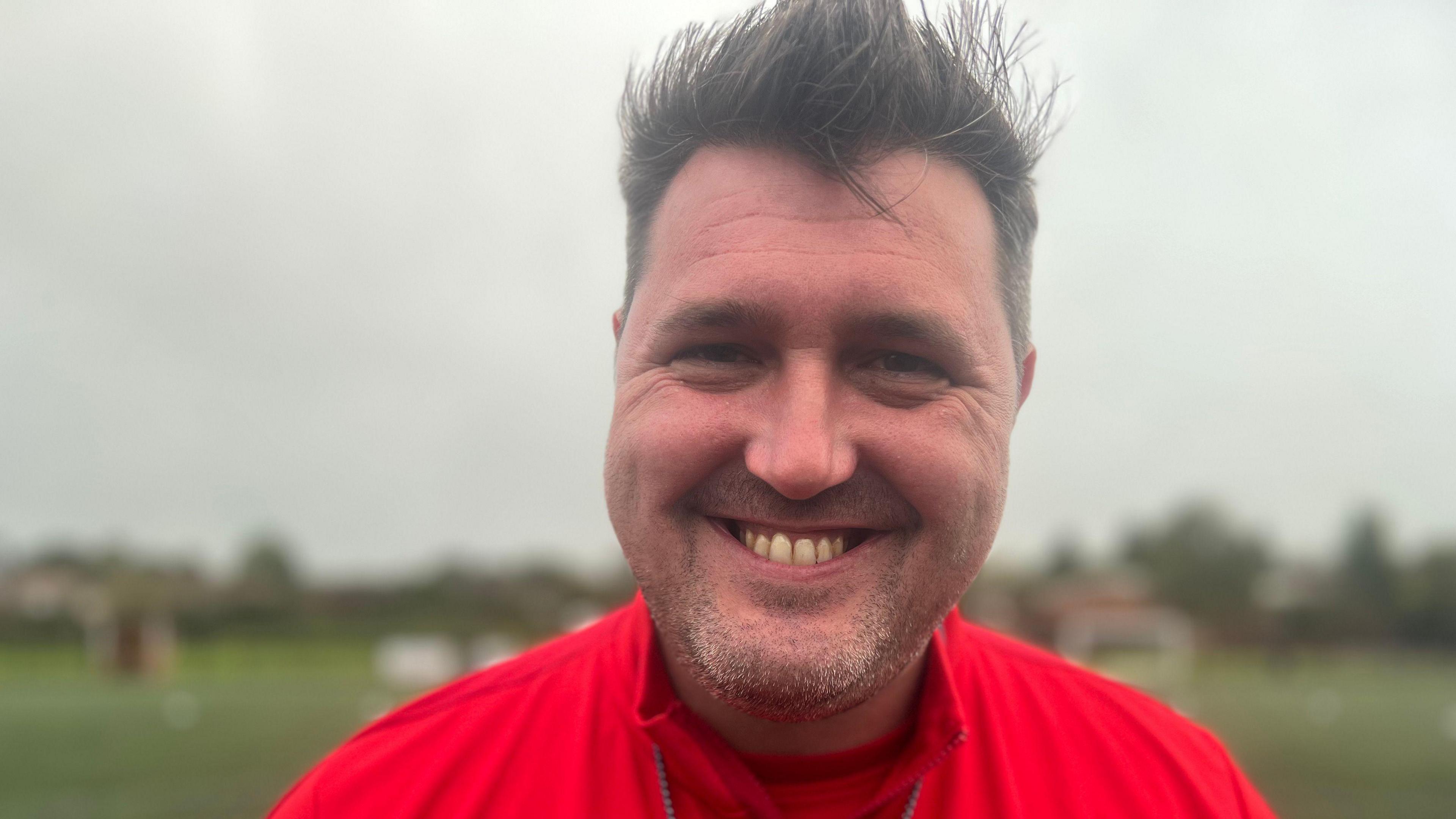 Nick Hawkins wearing red jacket and smiling at camera while standing on football pitch.