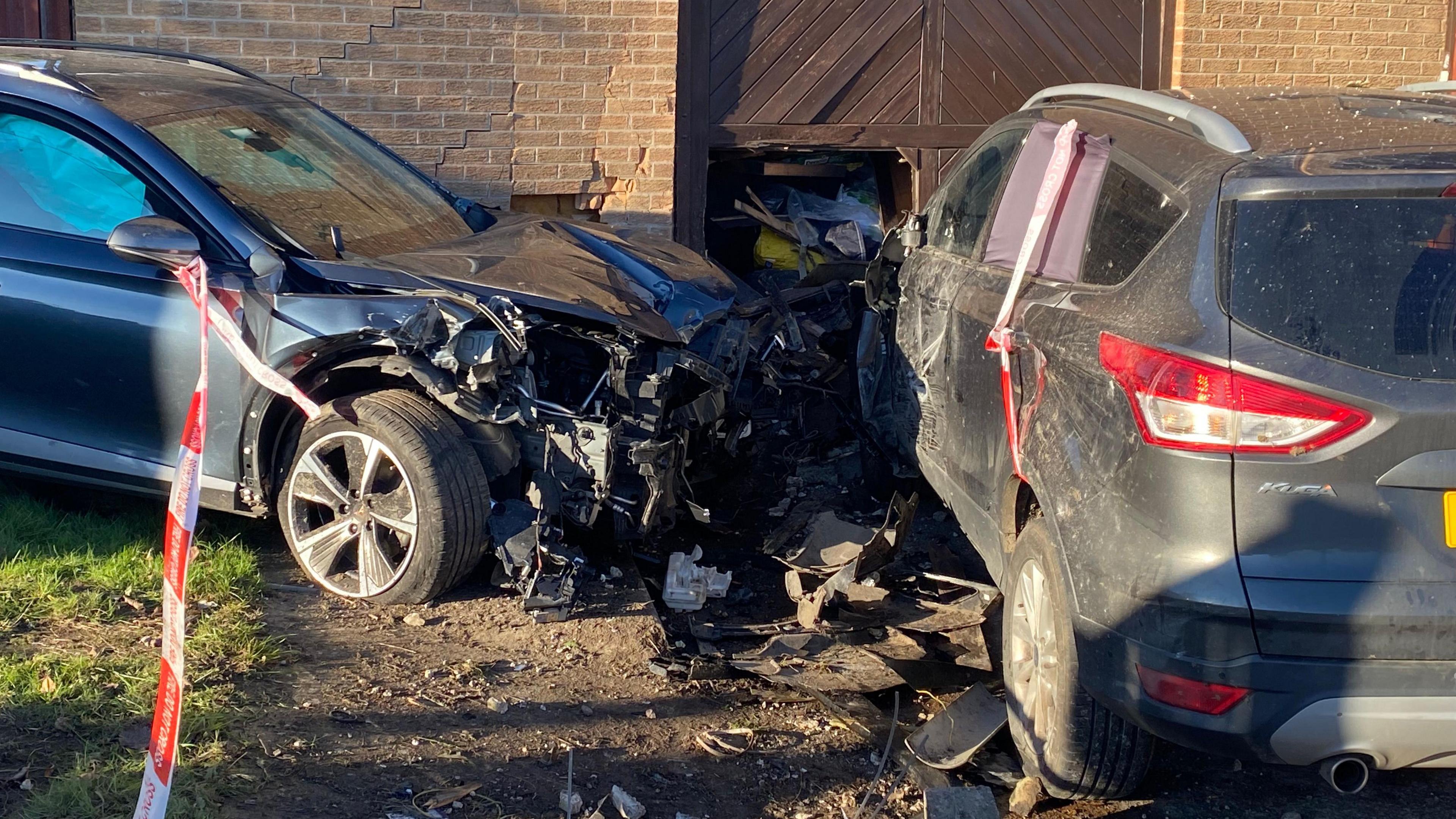 A car sitting parallel to a house. The wall of the property is cracked and has caved in. The front end of the car is crumpled and completely destroyed. The car's airbag has gone off and can be seen through one of the front windows.