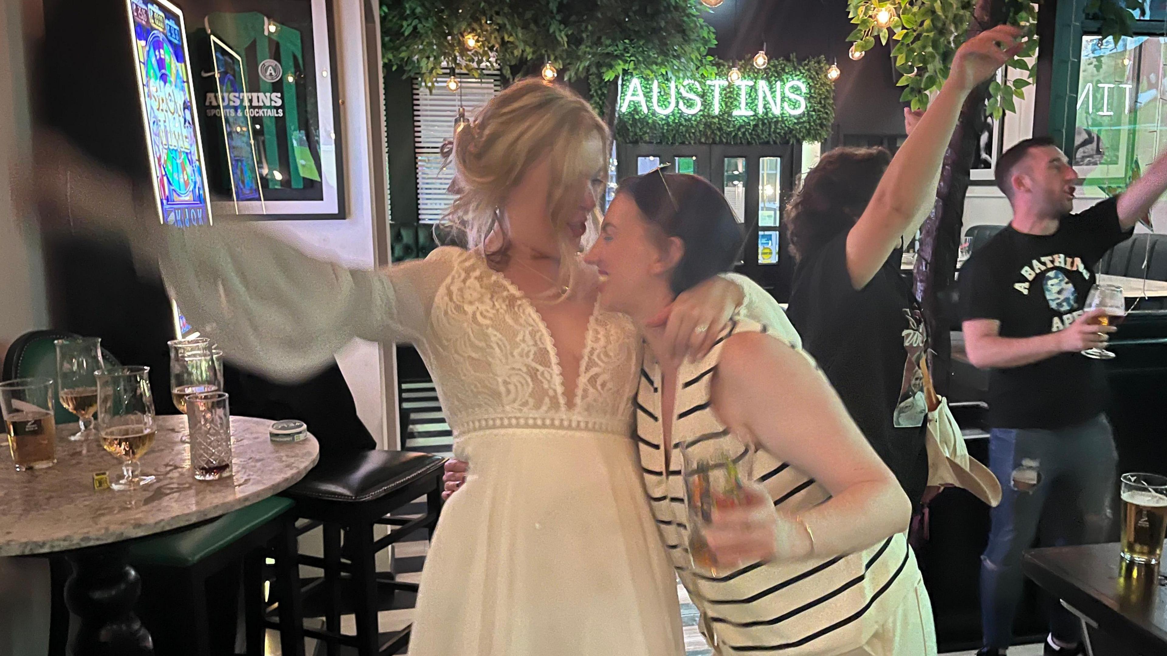A woman in a wedding dress is hugging a friend in a white and black striped top in a bar. Behind them, other people can be seen celebrating, tables are full of drinks, and there's a light-up sign says "AUSTINS"