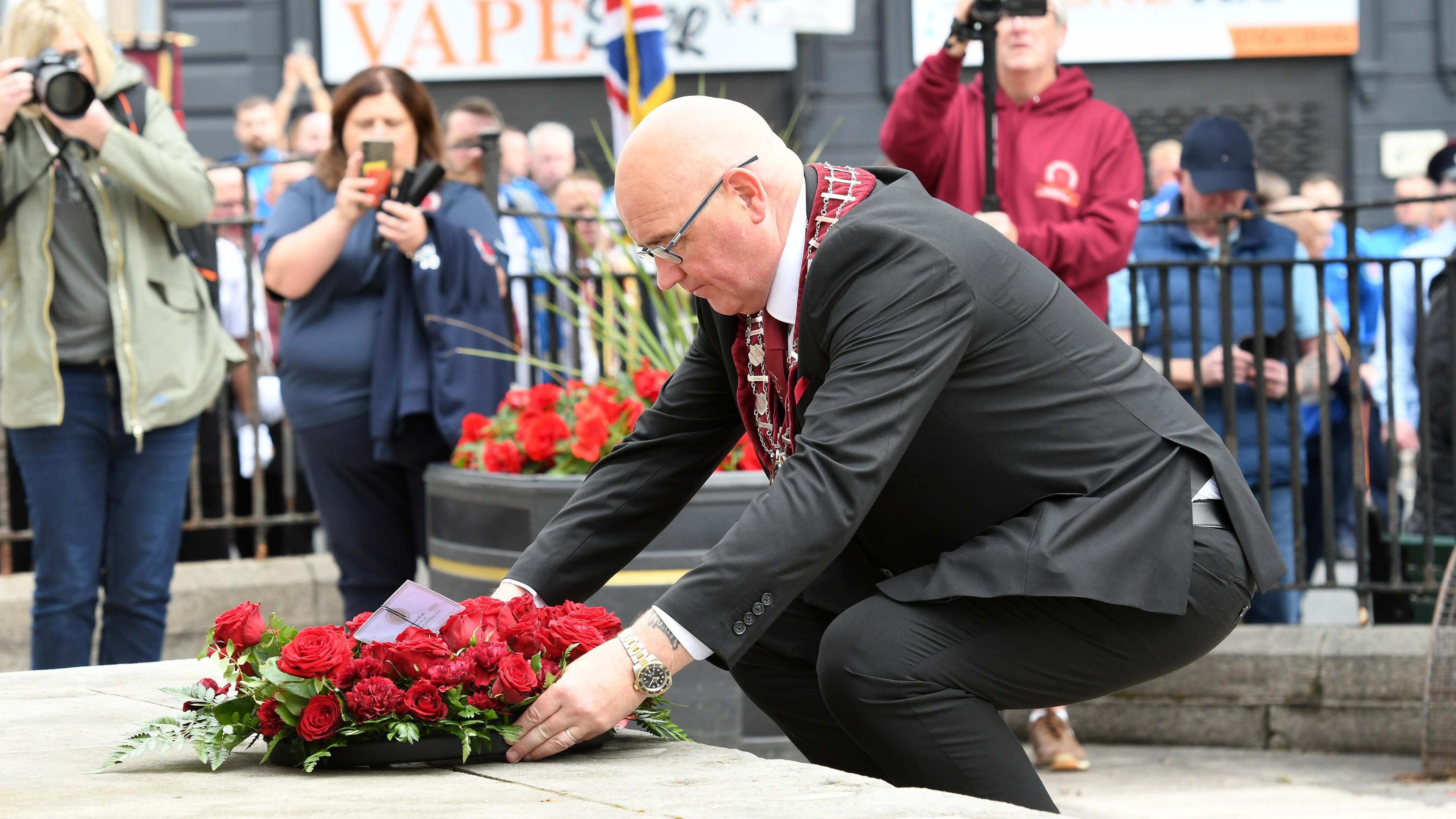 William Walker kneels to lay a wreath of red roses. He is wearing a suit and a maroon sash. There are three people in the background, taking pictures or filming