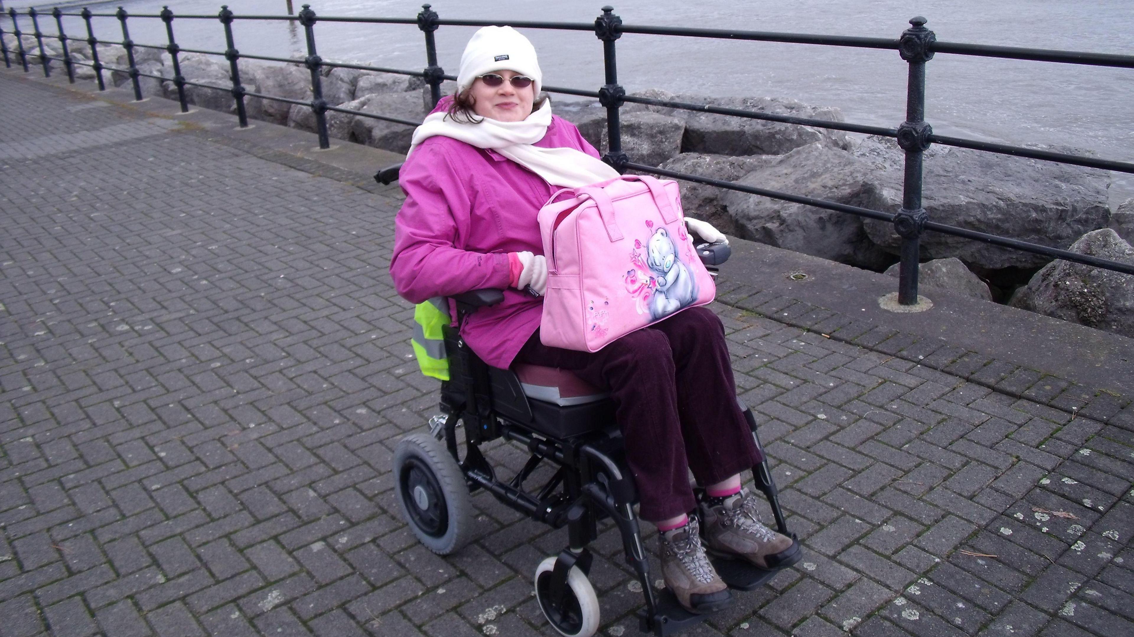 A woman sits in a wheelchair wrapped up warm with a pink coat and a white hat and scarf. She is sitting above a number of rocks at the seaside