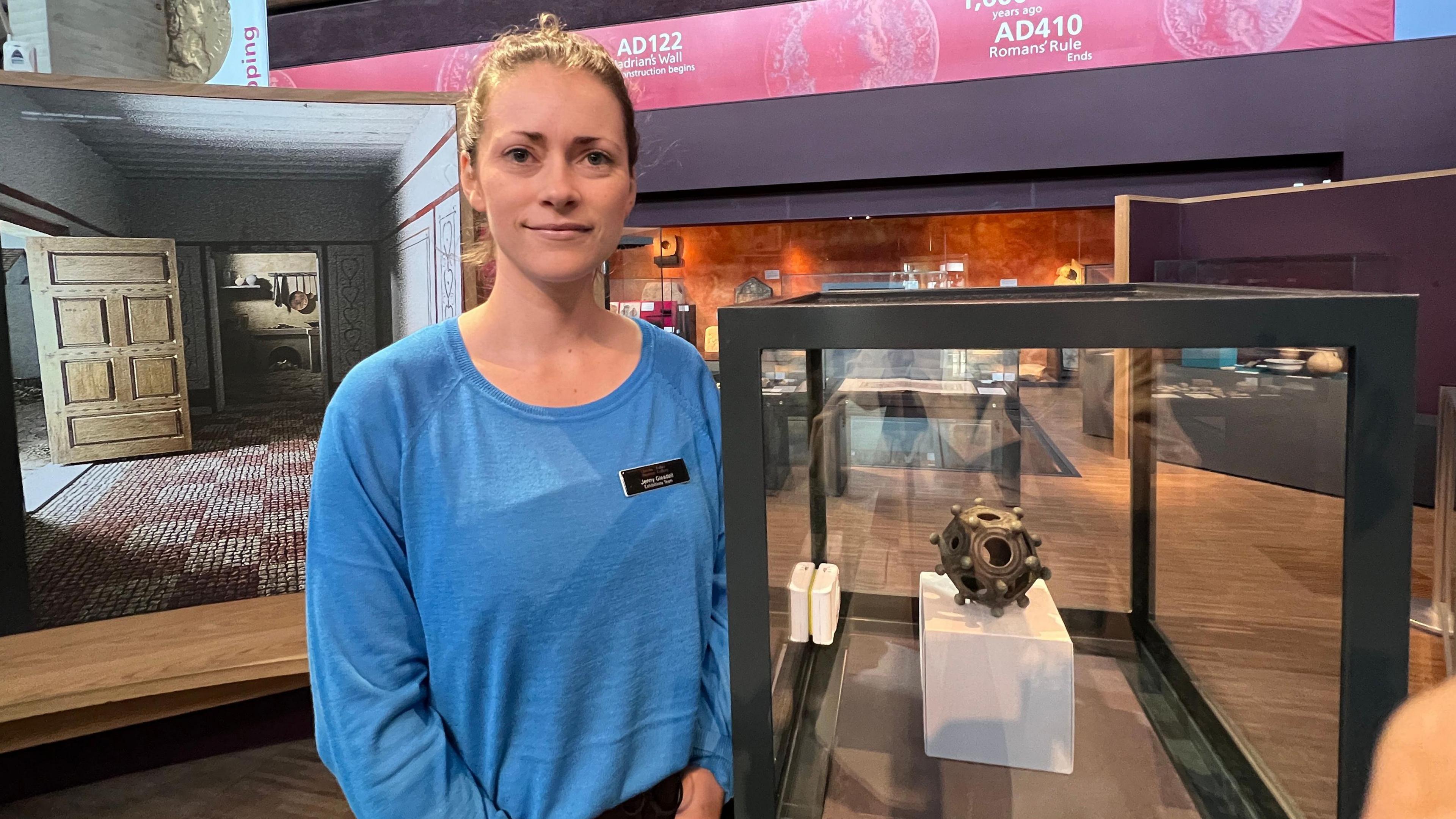 Jenny Gleadell wearing a blue top with name badge standing next to a black-rimmed glass display case containing the mysterious artefact 