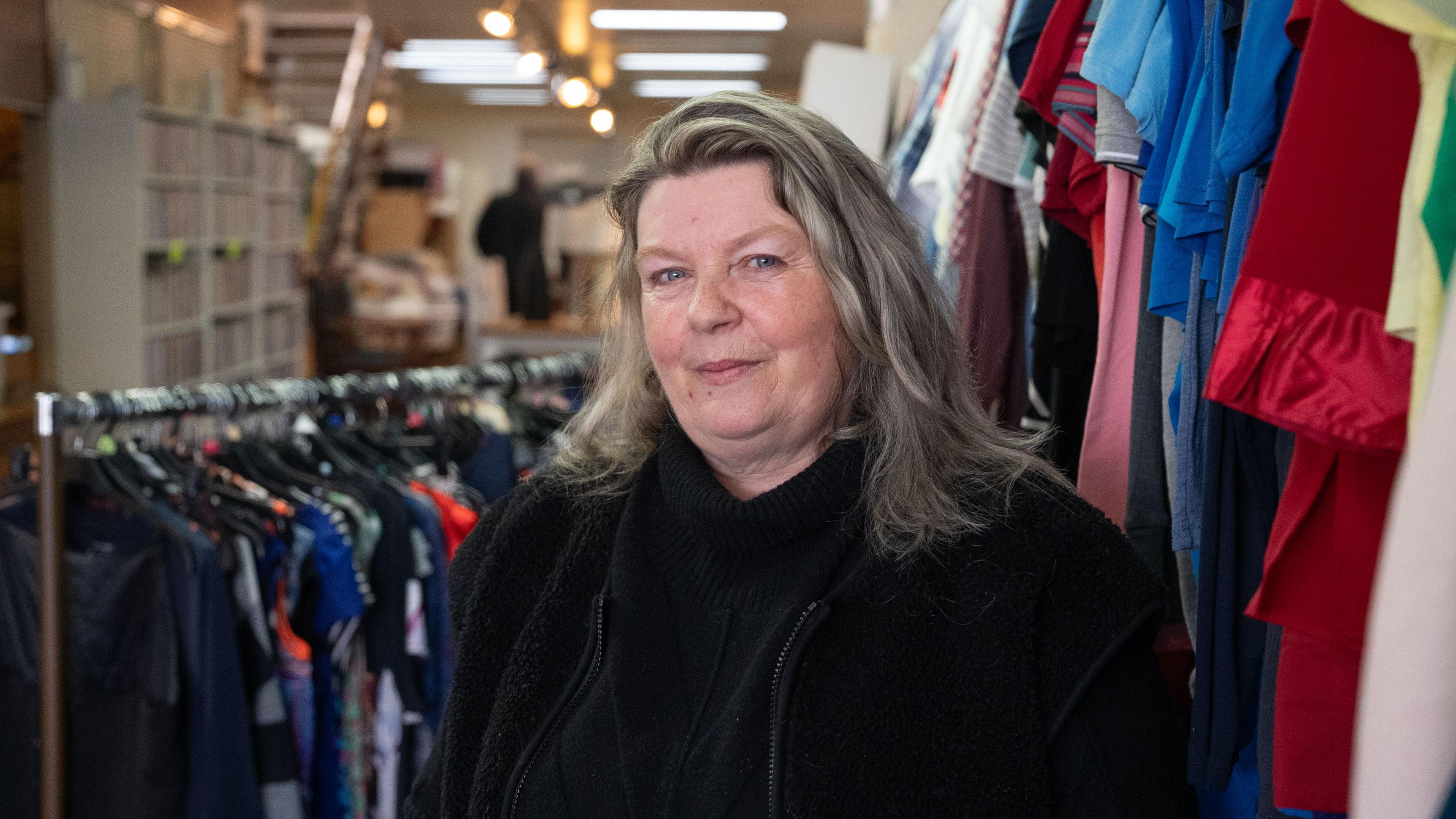 A woman with grey hair swept back over her head looks at the camera. she is wearing a black fleece and top. Behind her are clothes racks and shelves of goods. 