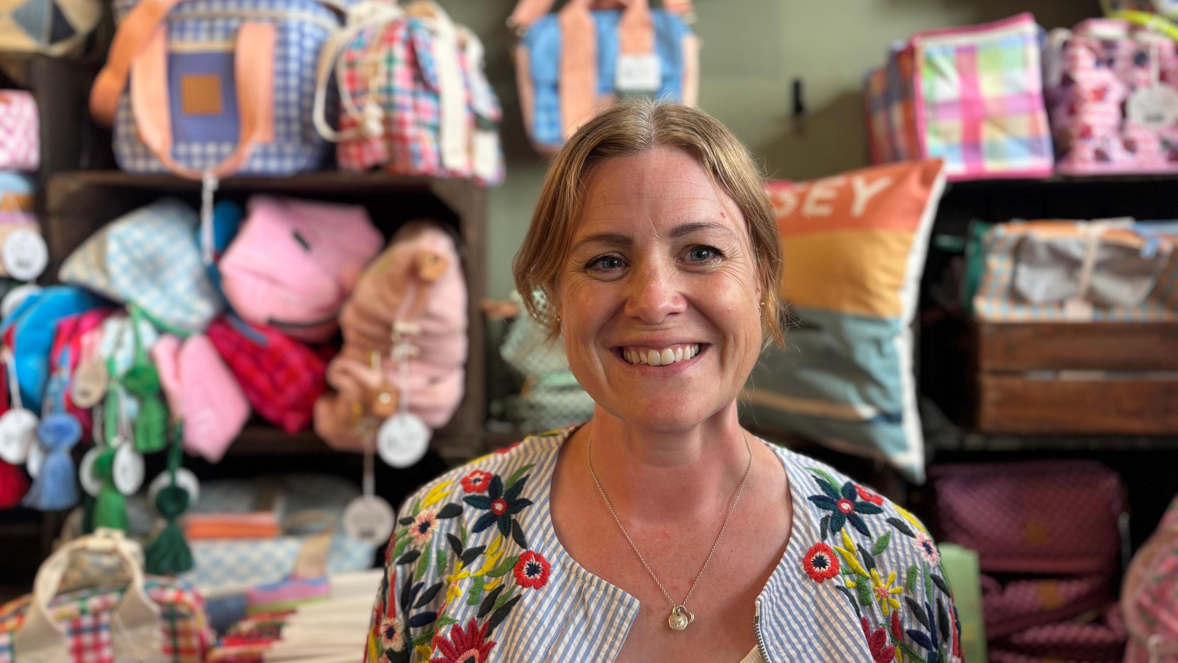 Harriet Rouse smiling towards the camera in her shop. She is wearing a stripy and floral jacket and is wearing a necklace. In the background there are colourful items on shelves. 