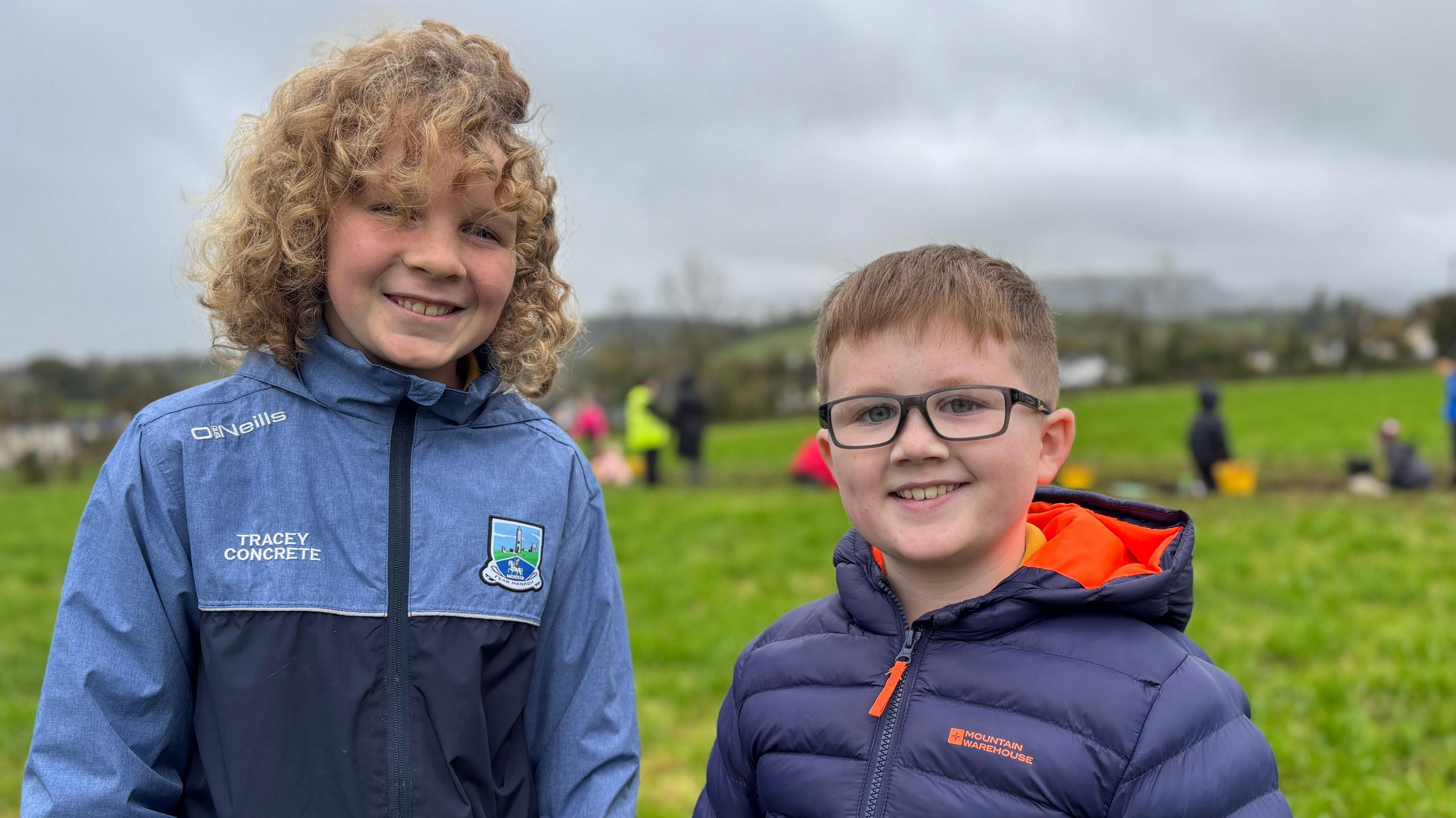 Two boys stand in a field. They are warmly dressed. The taller boy has blond curly hair and a big smile. The boy beside him has a wide smile and is wearing glasses. Behind them is a field with people and buckets. 