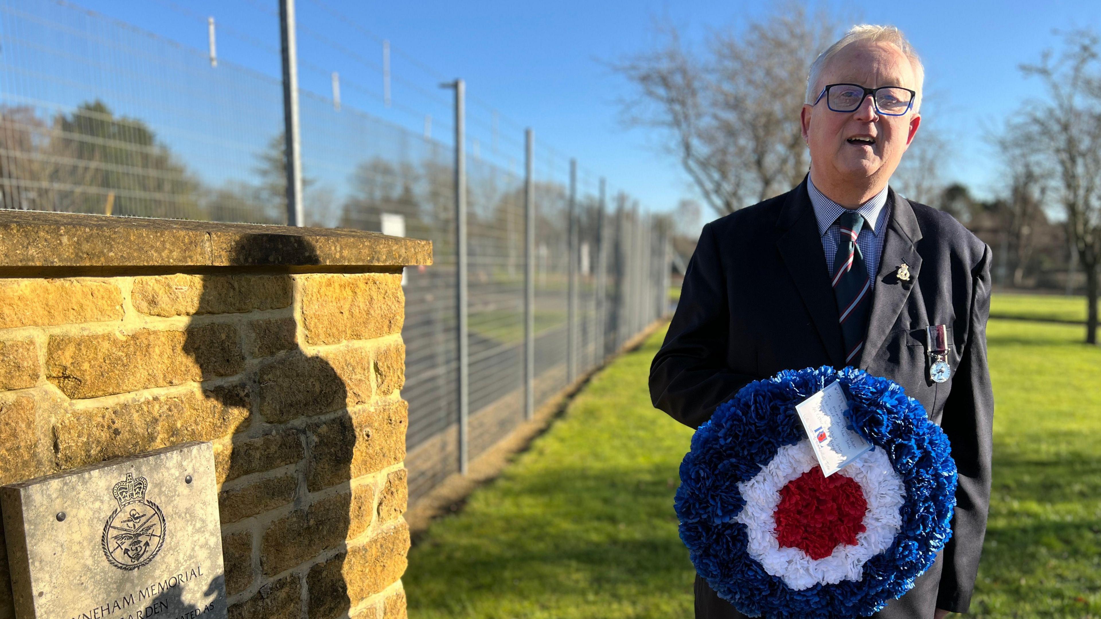 Geoff Bridgeman holding a memorial wreath. He is wearing a dark suit and is smiling into the camera.