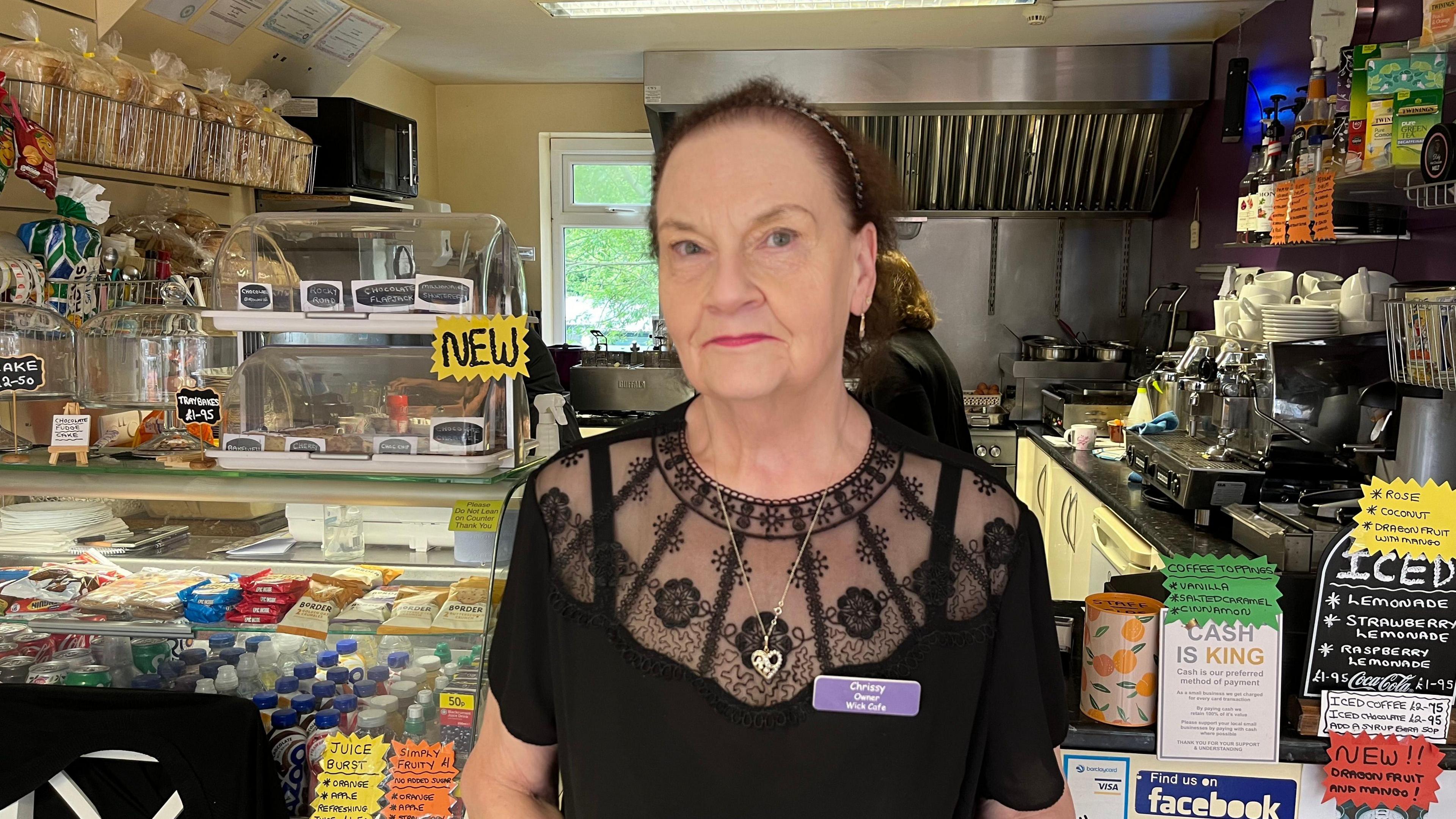 Woman with dark hair and magenta lipstick, wearing a purple name badge and standing in front of a cafe kiosk filled with drinks and snacks