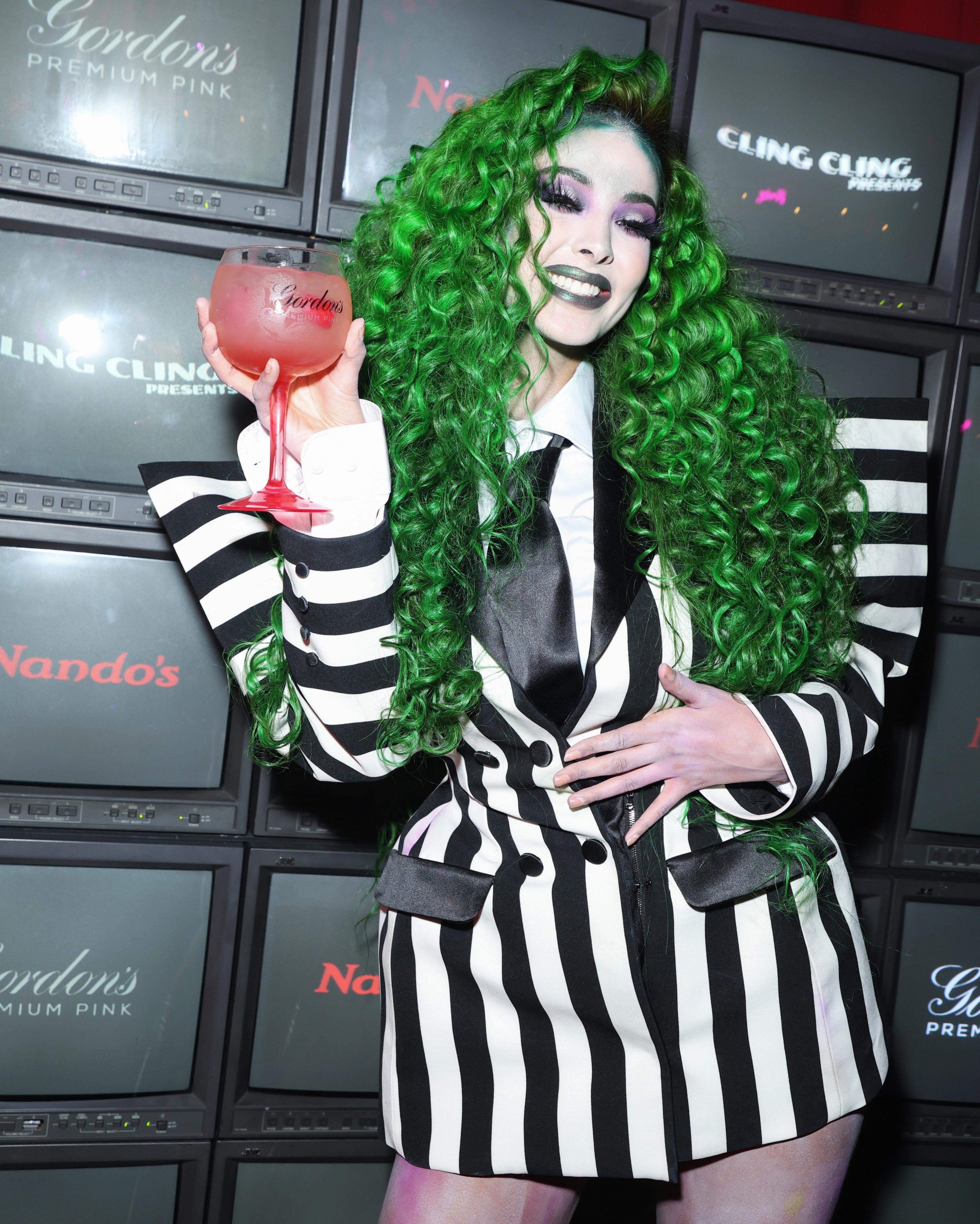 Maya Jama attends a Halloween party in a costume inspired by the title character from 1988 film Beetlejuice. She has long green hair and is wearing a blazer with wide-square shoulders and black and white stripes. 