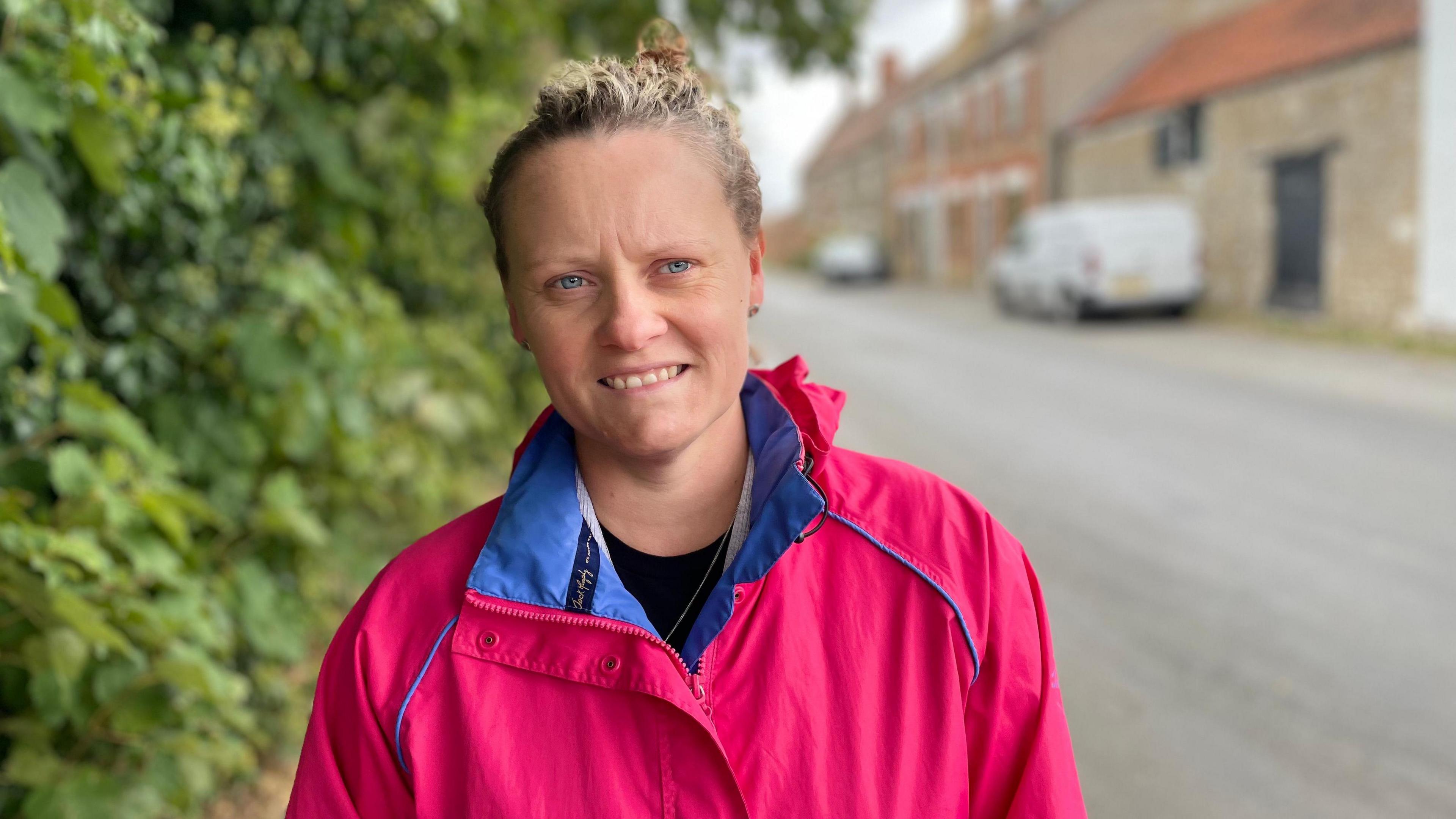 A woman in a pink raincoat is standing in the street in Rippingale and looking into the camera. A white van and red brick buildings are visible in the background.