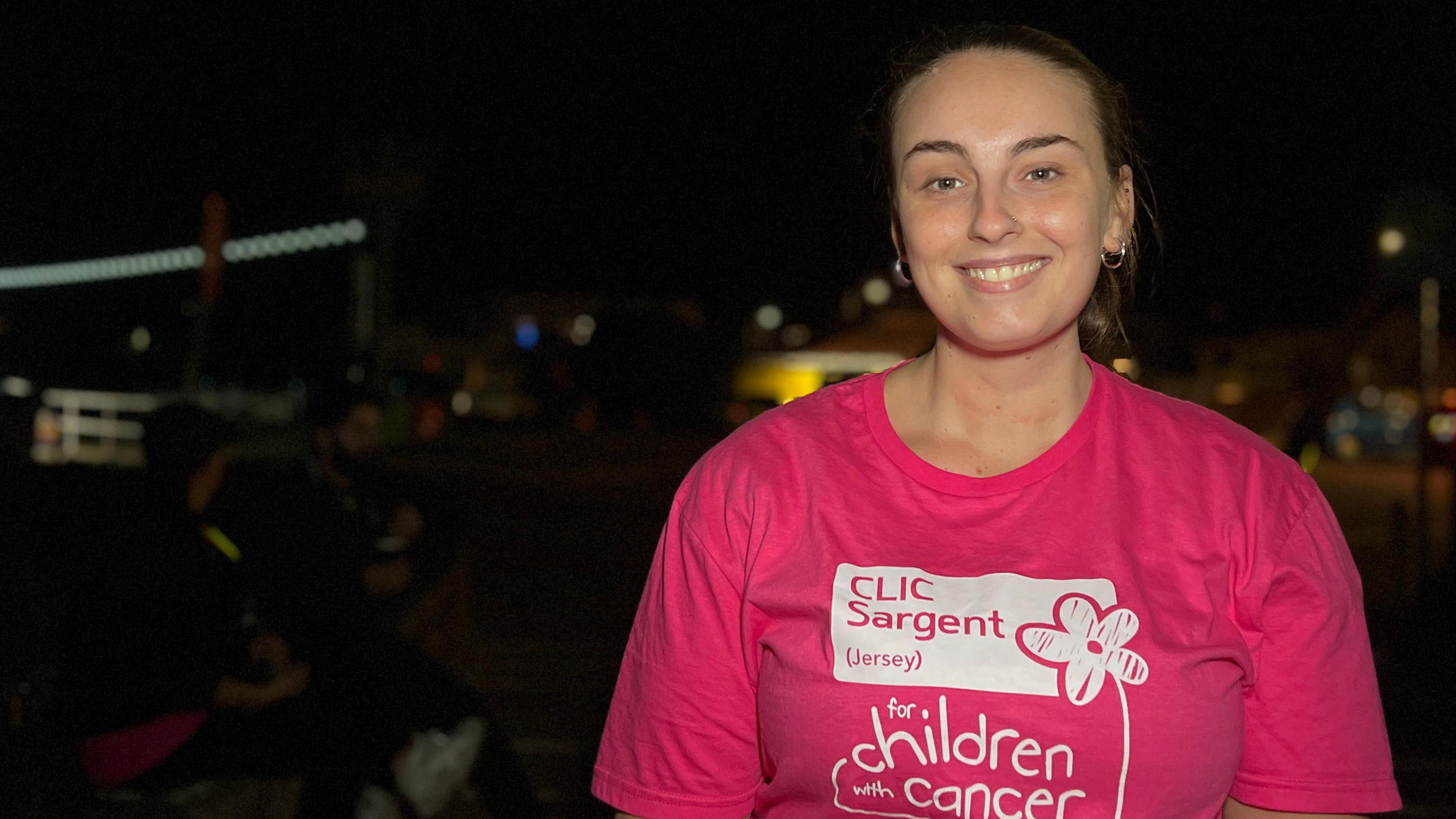 Antonia stands outdoors at night wearing a bright pink T-shirt that reads "CLIC Sargent (Jersey) for children with cancer." The background is dark with scattered lights and indistinct figures.