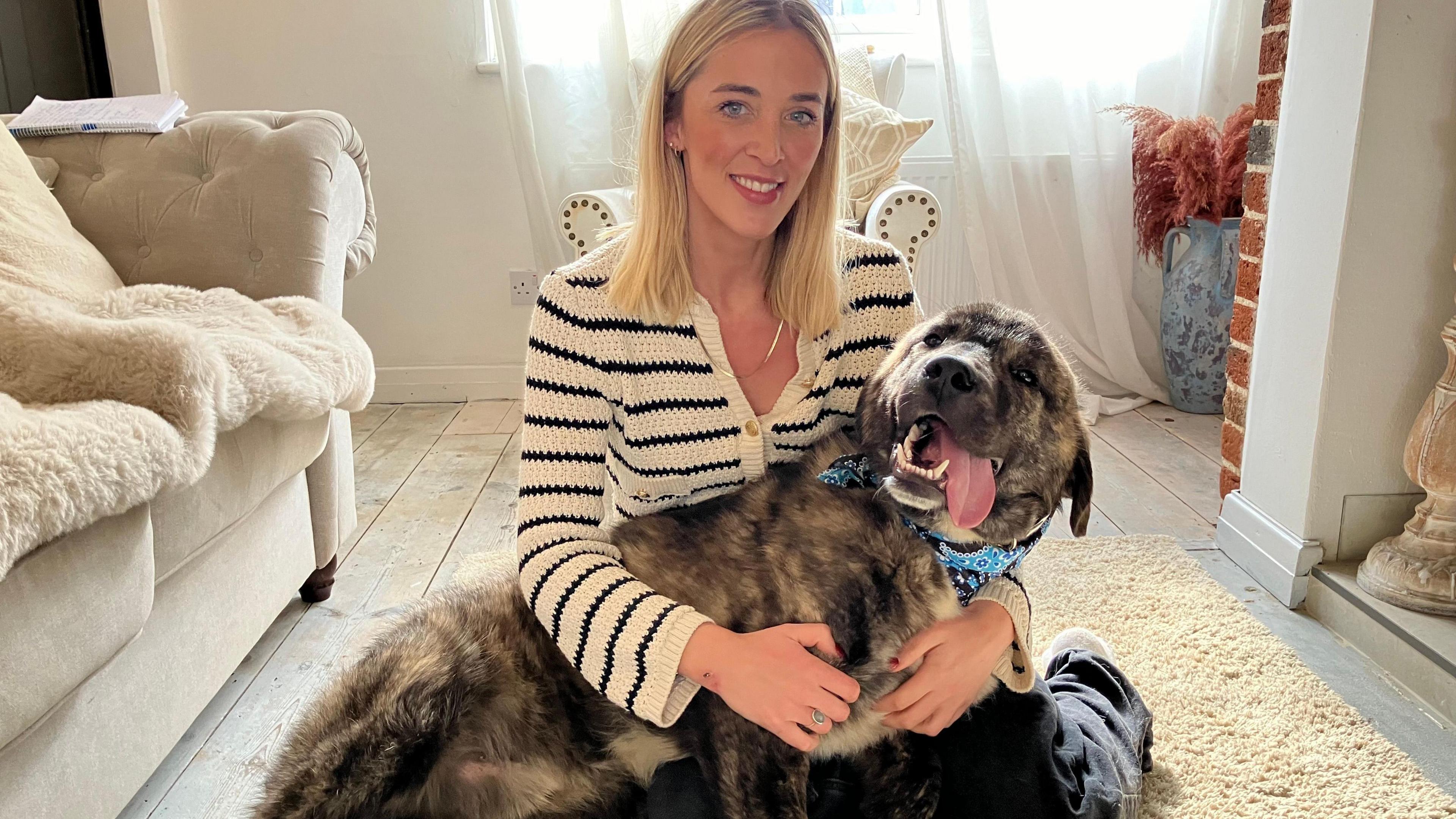 A dog with a cloth round his neck and his tongue out in the embrace of Felicity Sutton, who's blonde, is wearing a striped top and is smiling at the camera. They are sitting on the floor of a living room